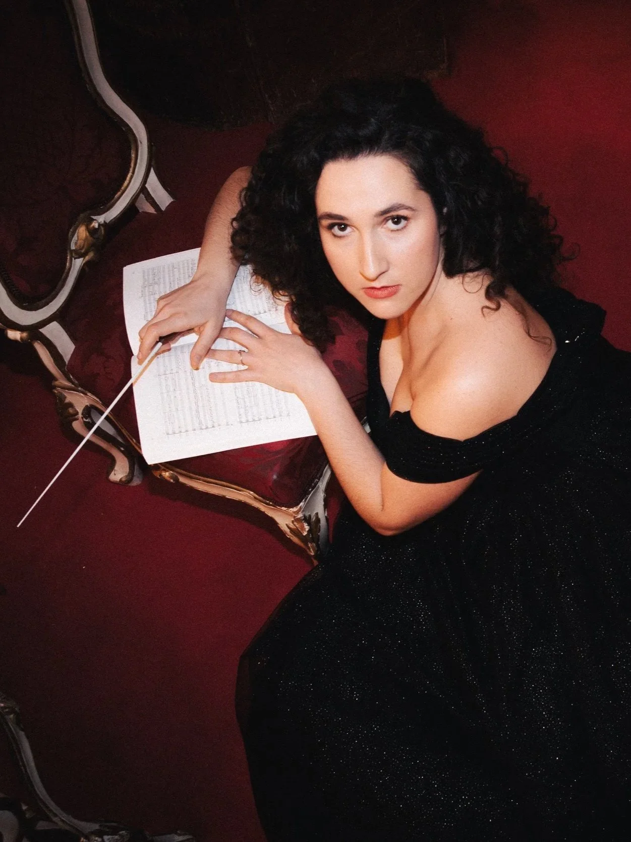 A woman with curly dark hair wearing a black dress, lying on her stomach on a red velvet antique chair, looking up into the camera while holding a conductor's baton and surrounded by sheet music.