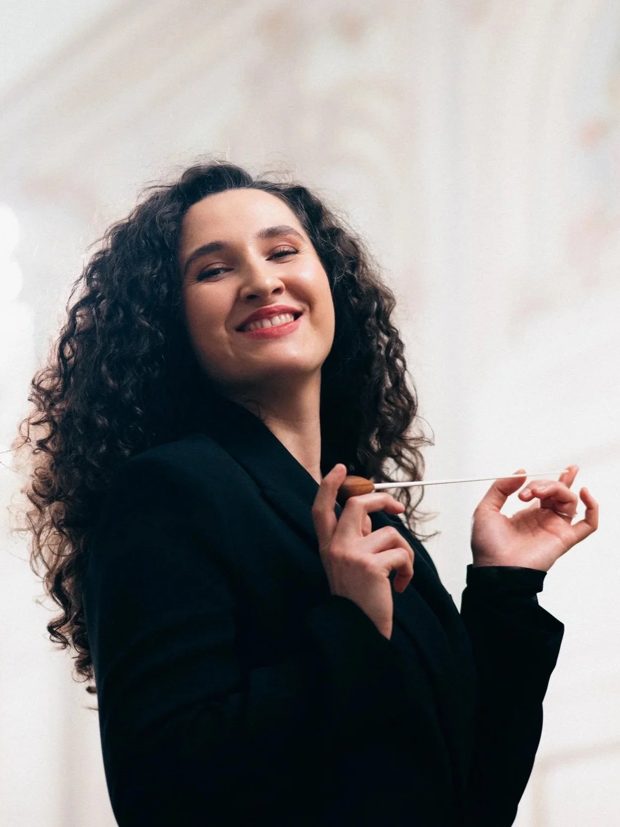 A conductor with curly hair smiling, dressed in black, holding a baton in her right hand against a plain white background.