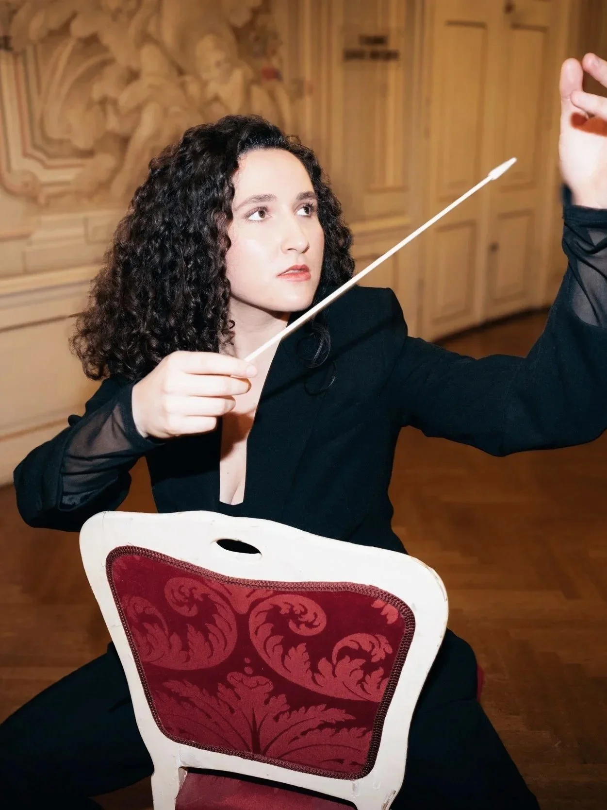 A woman with curly dark hair, wearing a black outfit, is sitting on a chair with a red and black ornate pattern. She is holding a white conductor's baton and appears to be conducting, with her gaze focused upward. The background features a wooden wal