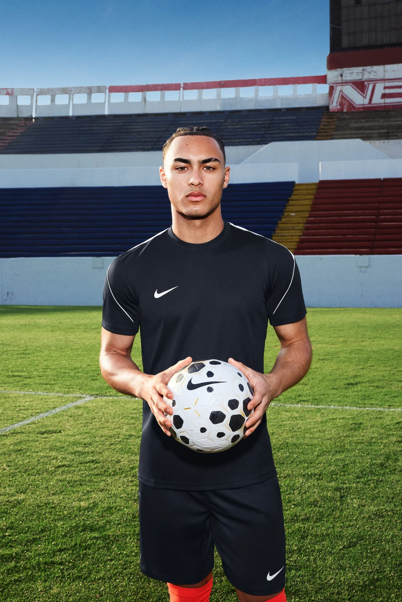 Young male soccer player in a black Nike training kit holding a soccer ball, standing on a pitch in front of empty stadium seating.