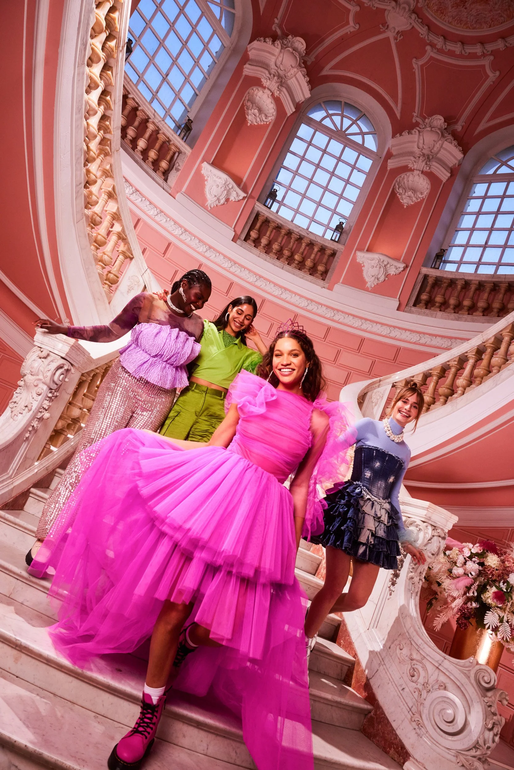 Four women in colorful evening dresses walking down an ornate pink staircase, laughing together.