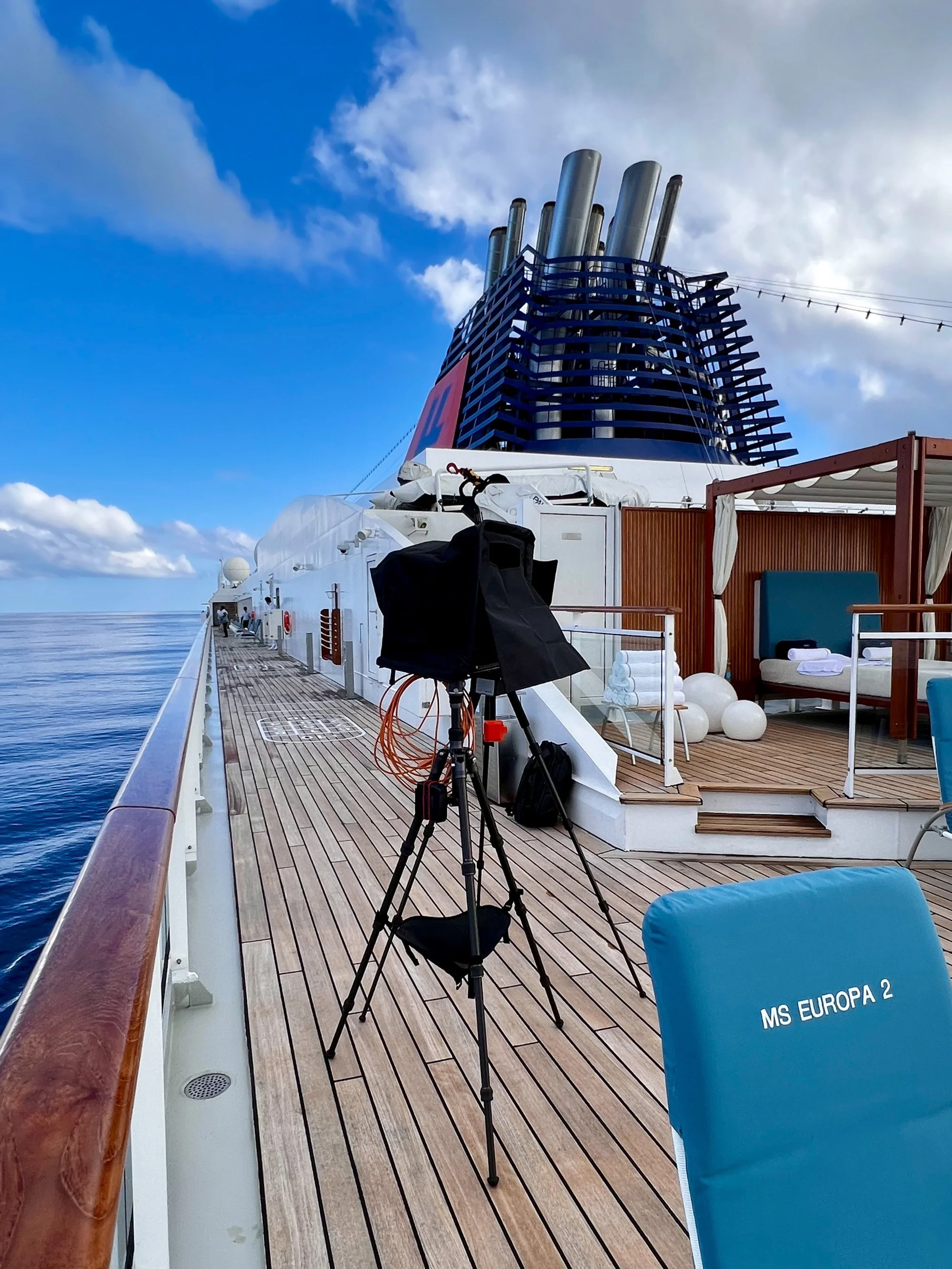 Photographer's monitor cart on a ship deck with a hooded screen on a tripod, funnel stacks and cabana seating in the background, labeled "MS Europa 2."