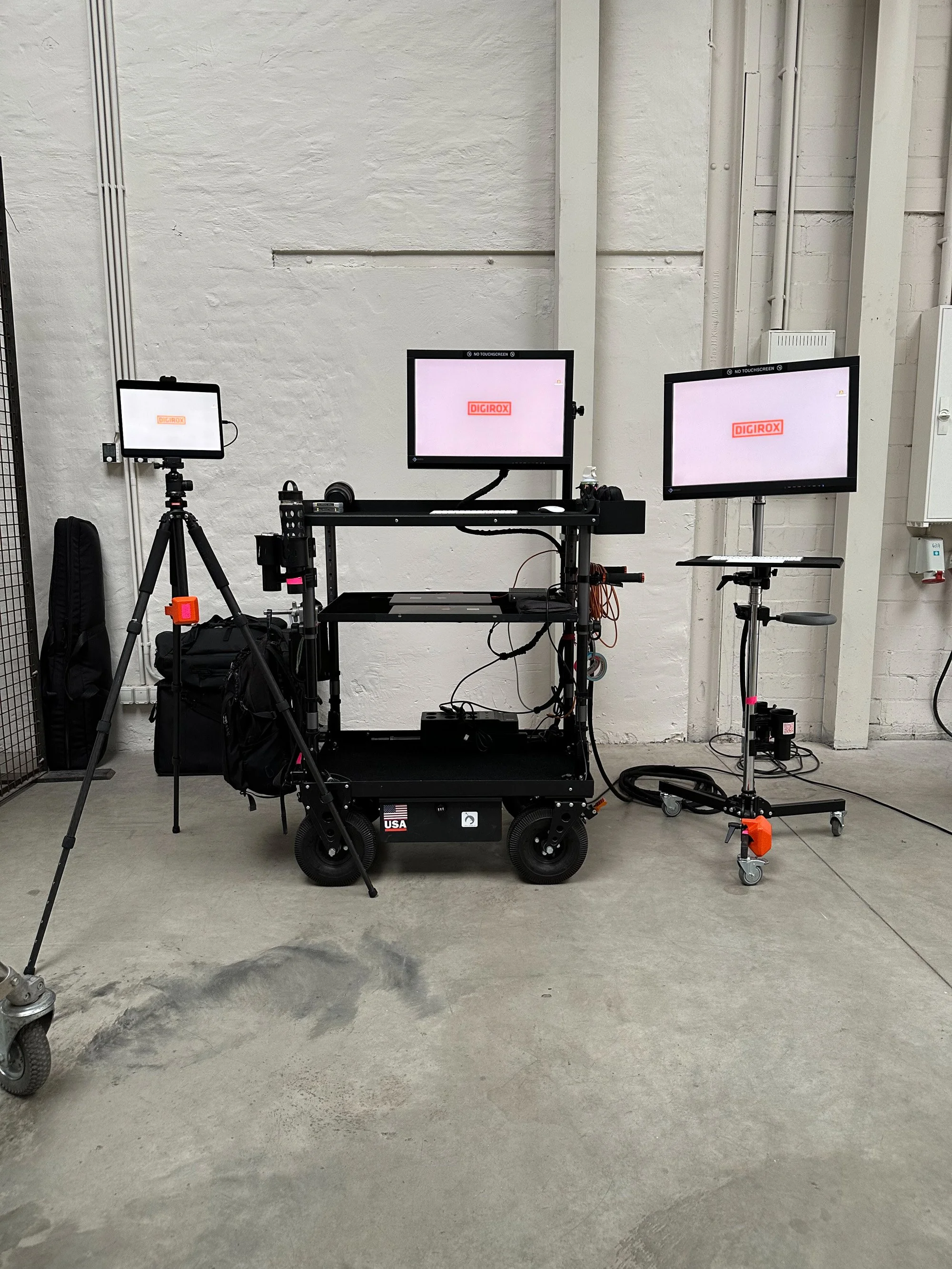 Minimalist studio setup against a bare wall with three monitors on stands displaying the DIGIROX logo, connected to a wheeled equipment cart.