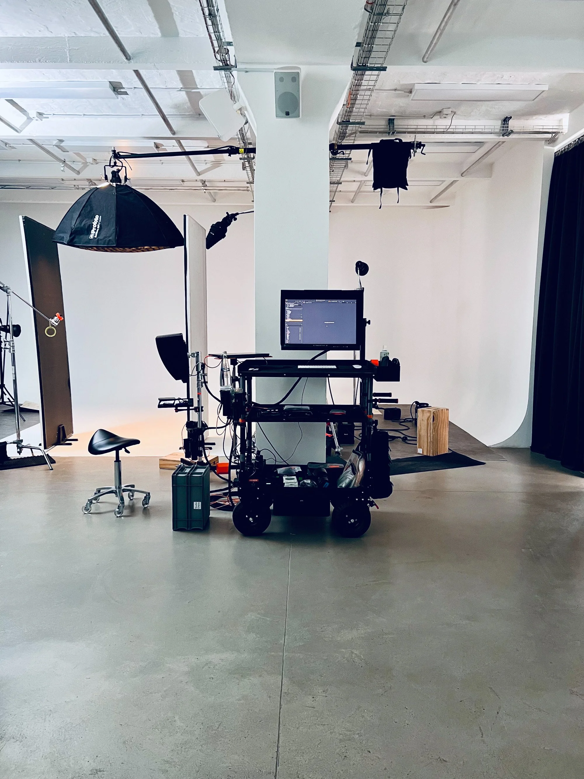 Photo studio with a white cyclorama backdrop, a softbox light overhead, and a monitor cart positioned beside a rolling stool.