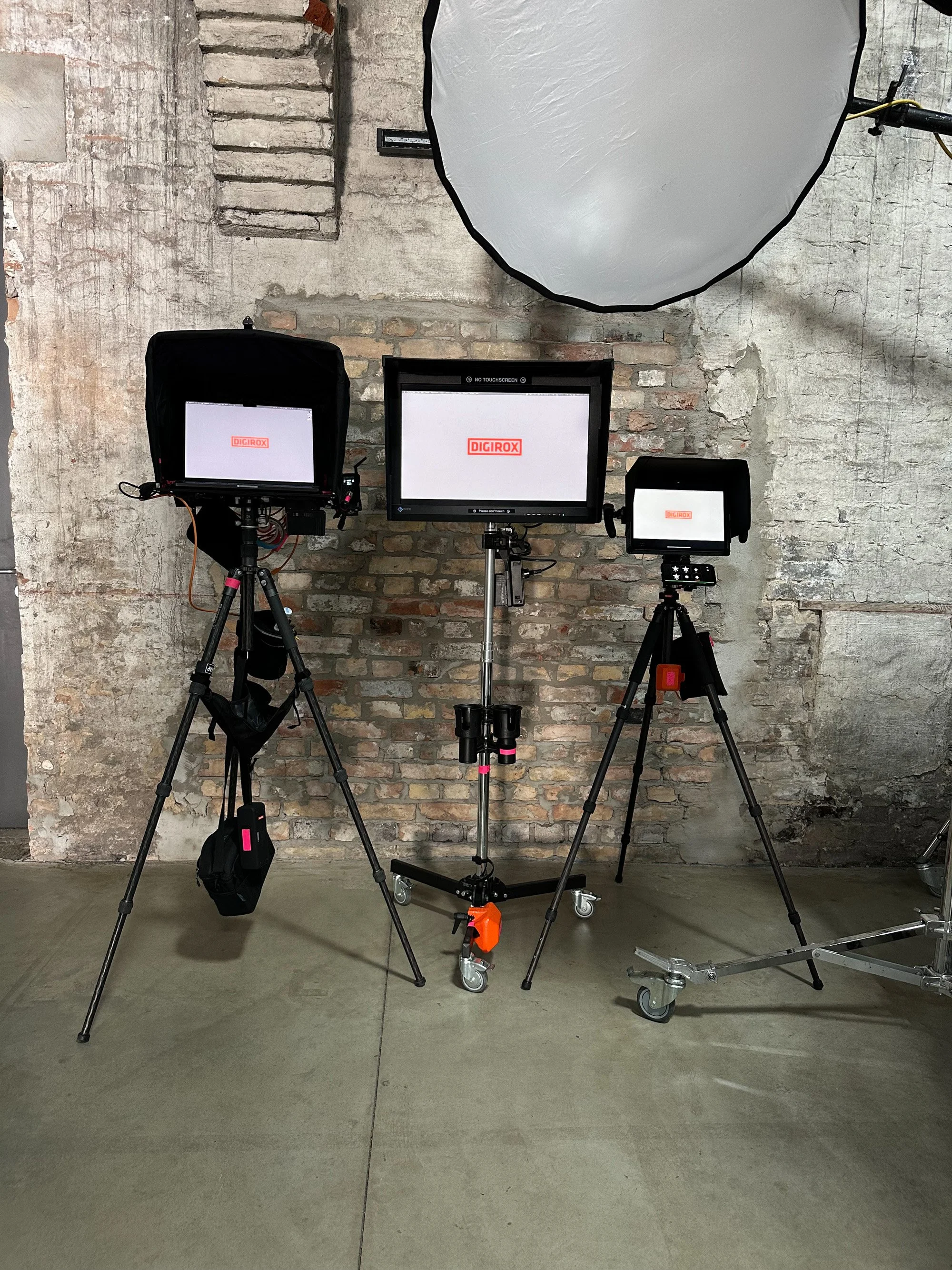 Three DIGIROX monitors on tripods against an exposed brick wall, with a large round reflector positioned above.