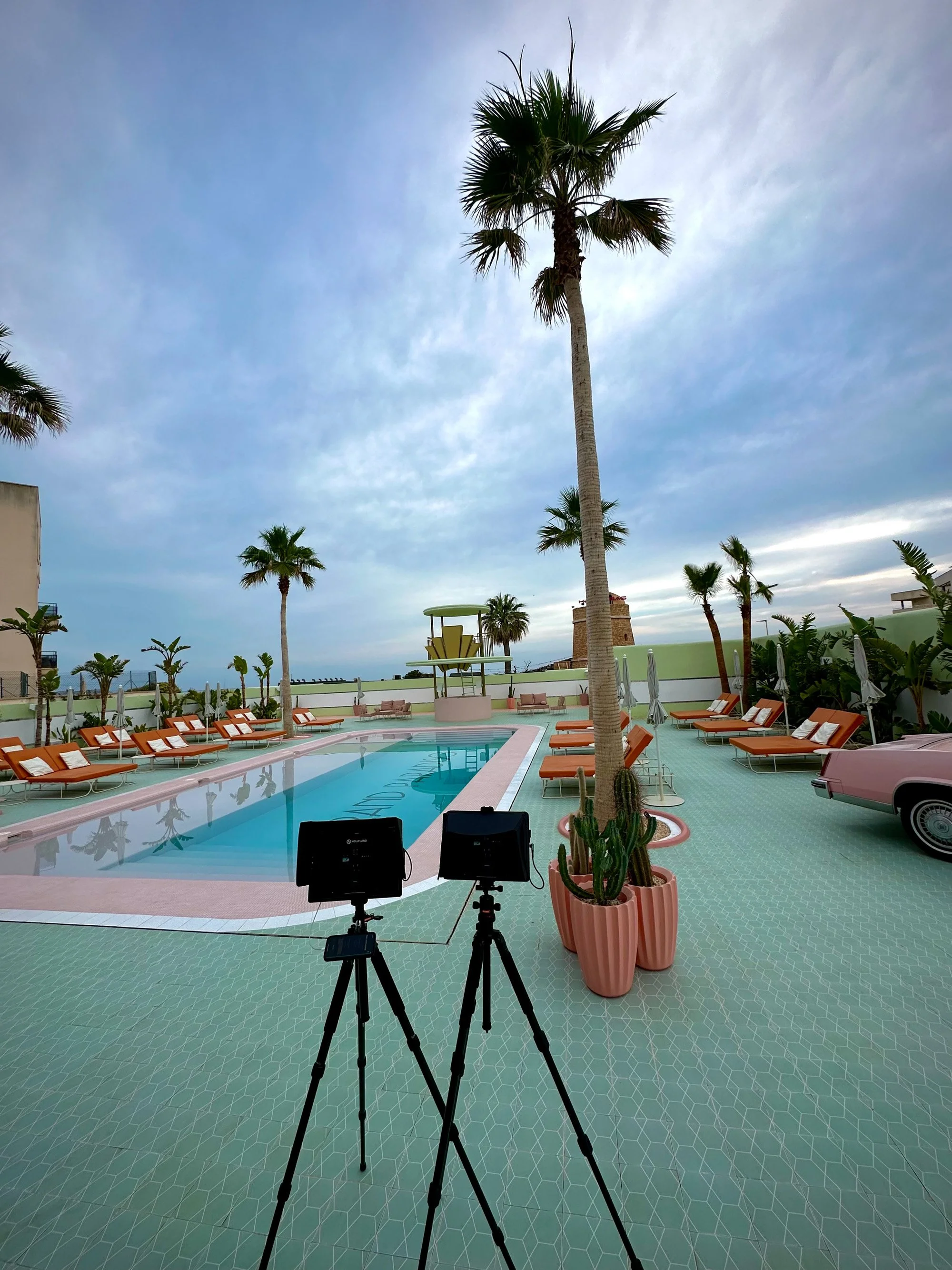 Two monitors on tripods set up on a pool deck with palm trees, pink tile flooring, and a vintage pink car nearby.