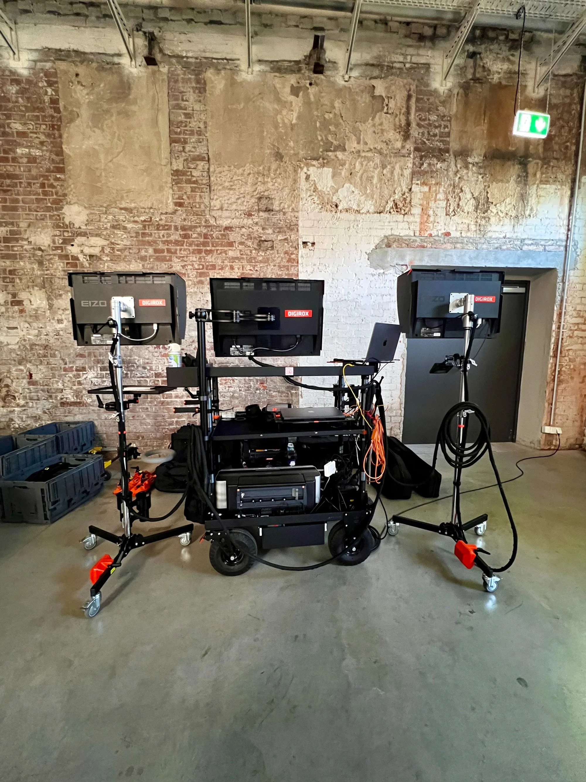 DIGIROX video village setup against a distressed brick wall, with three EIZO monitors, cables, and equipment crates.