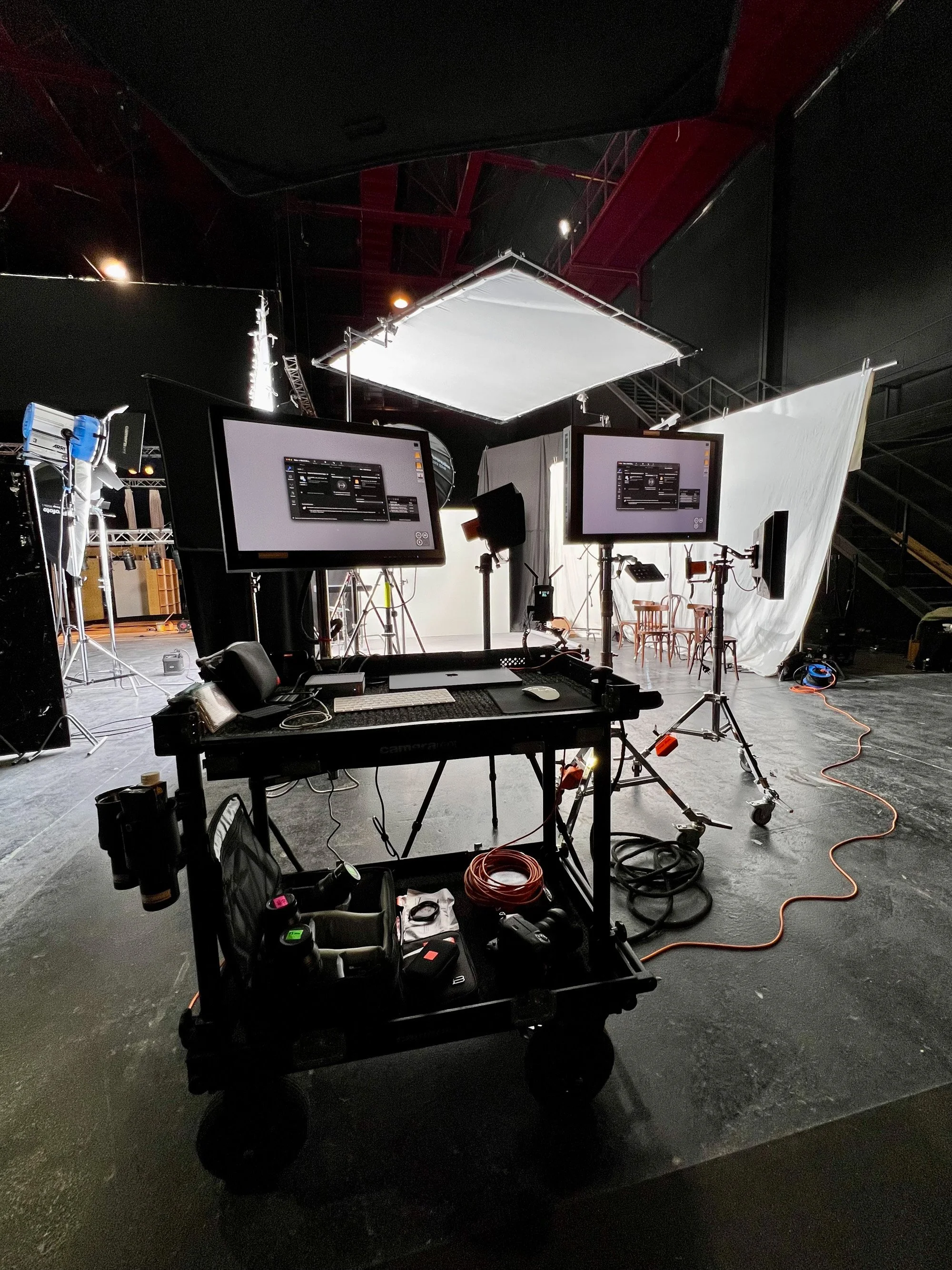 Monitor cart with two screens set up in a dark studio with red steel beams and a white bounce overhead.