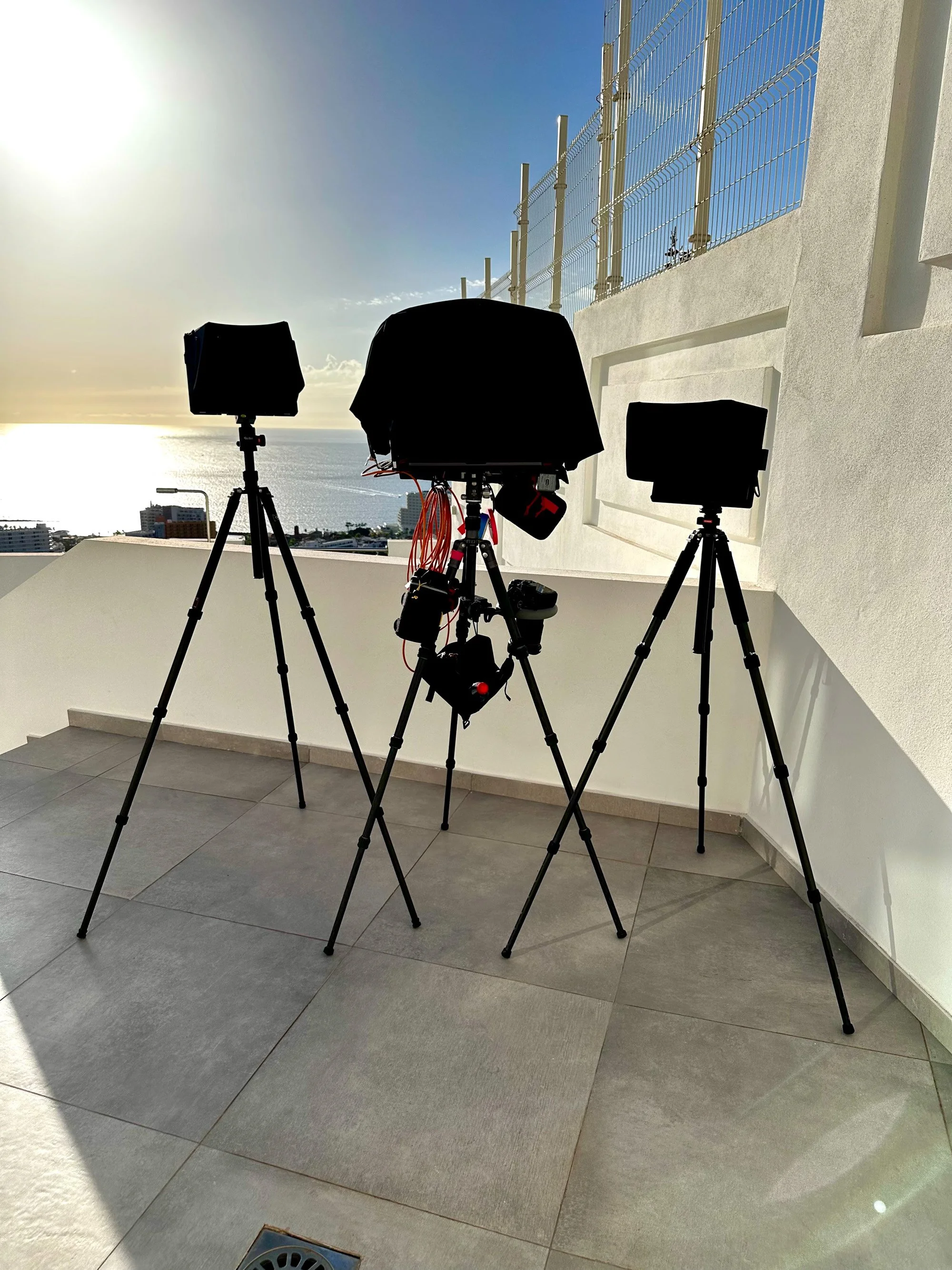 Three hooded monitors on tripods on a rooftop terrace overlooking the ocean at sunset.