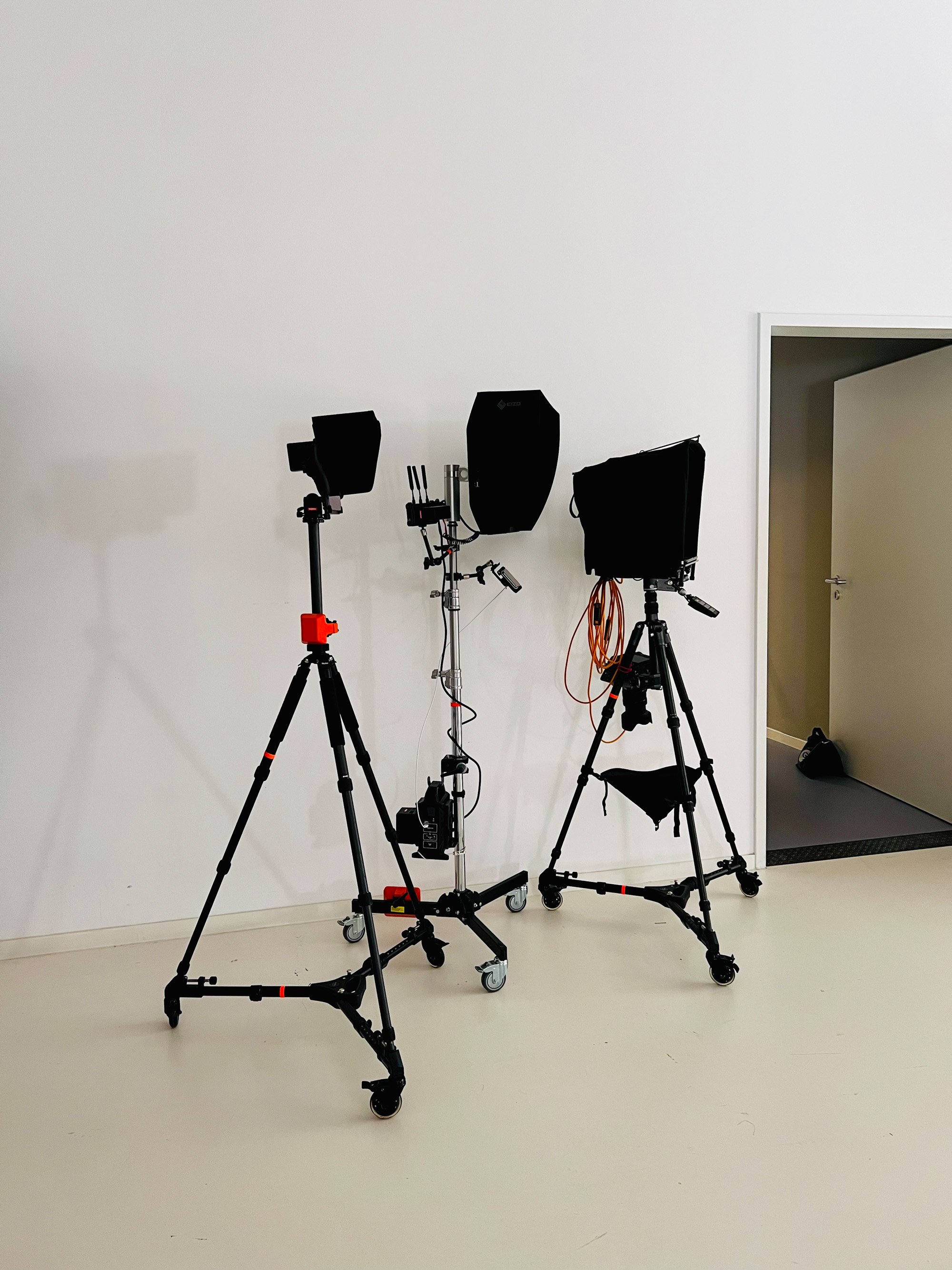 Three monitors on tripods in an empty white studio room, with a doorway leading to a blue-carpeted room.