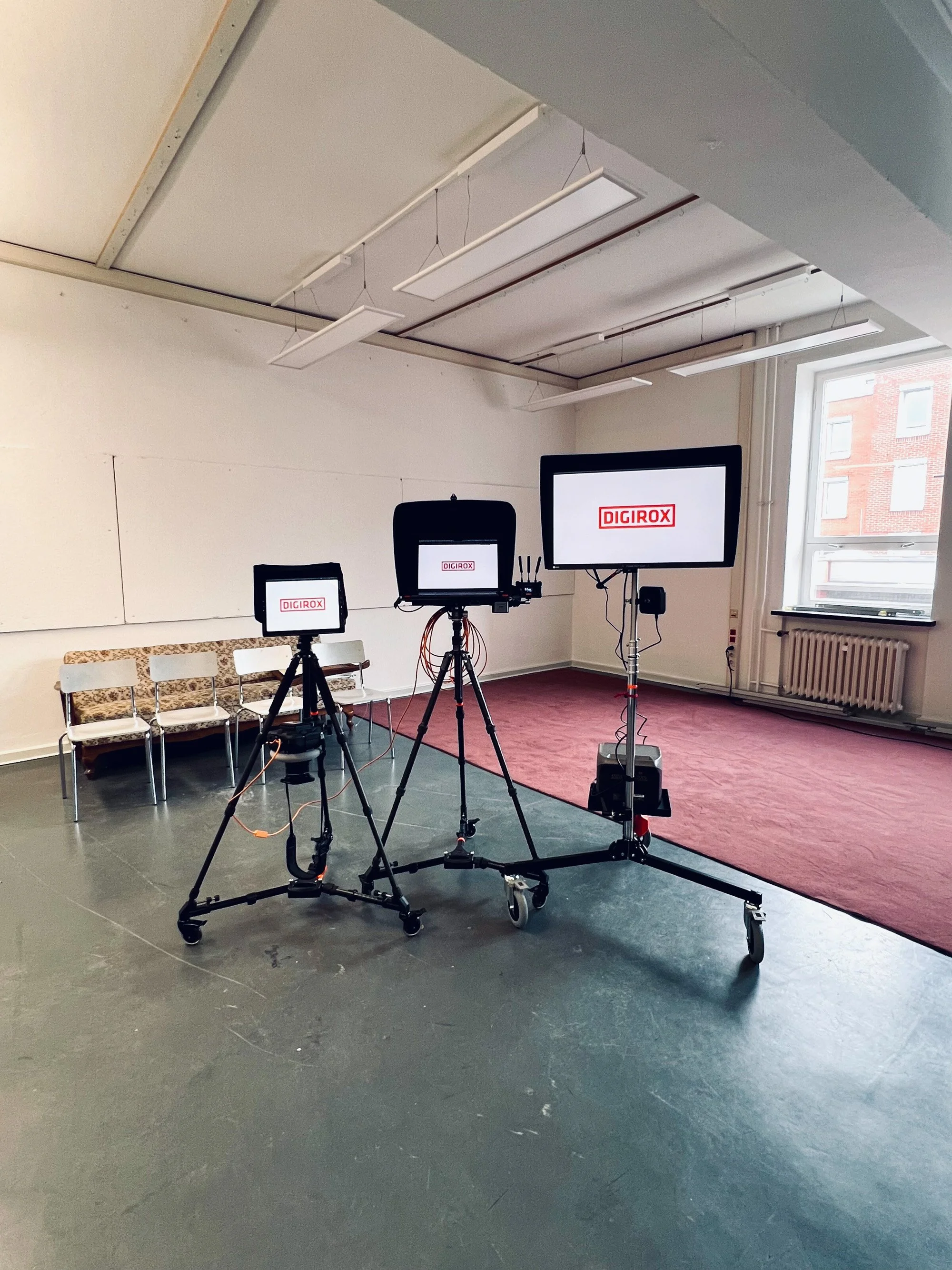 Three DIGIROX monitors on tripods in a room with a vintage bench, mismatched chairs, and maroon carpet.