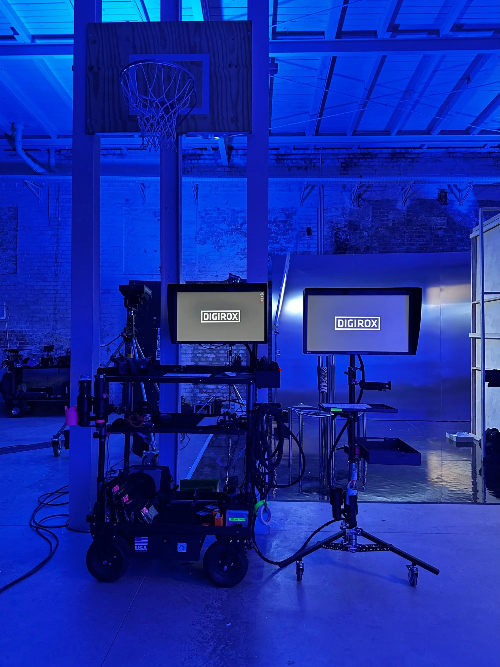 Two DIGIROX monitor carts in an industrial space lit in blue, with a basketball hoop and mirrored backdrop panel.