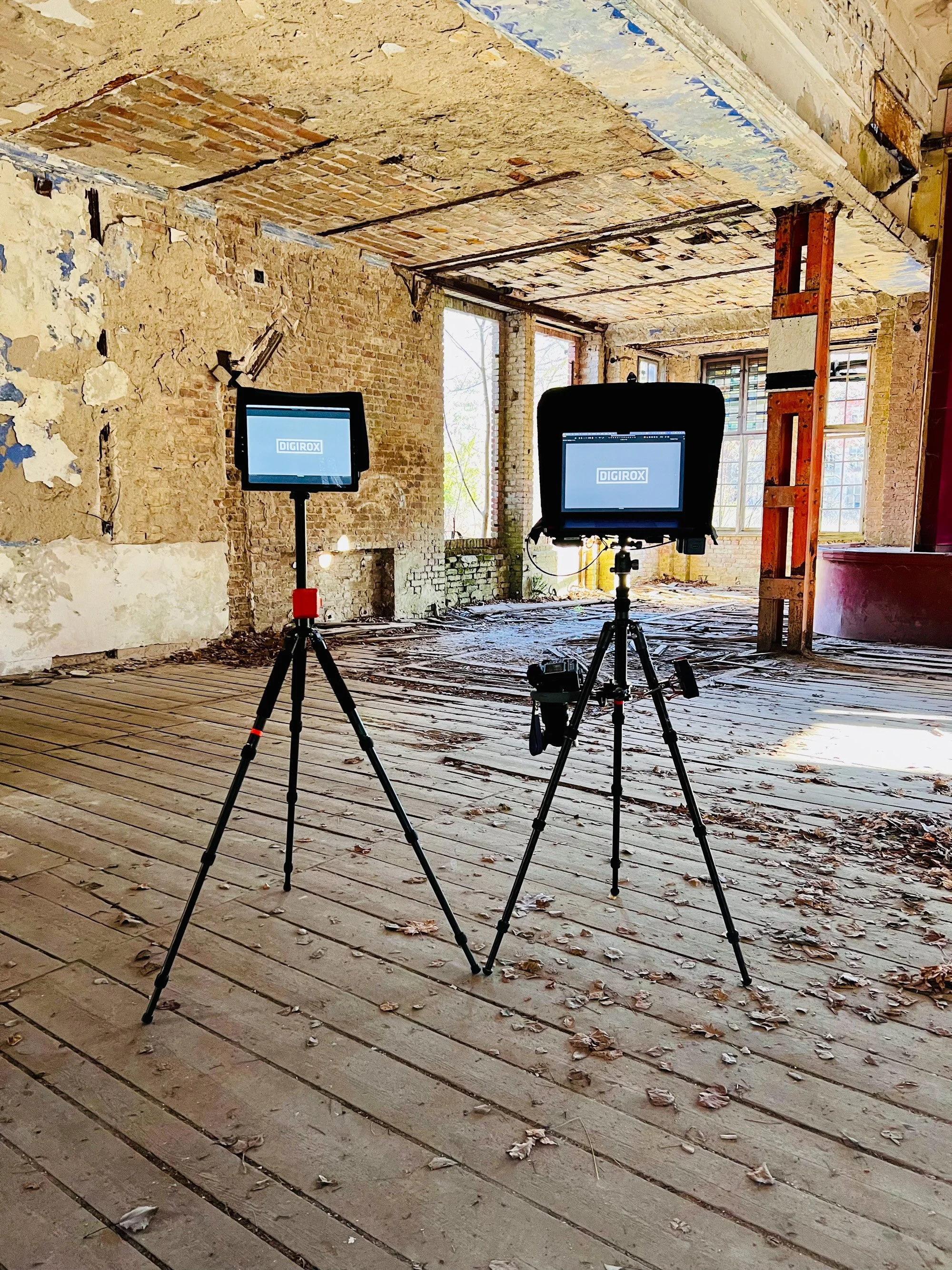 Two monitors on tripods in a derelict building interior, with brick walls, a distressed ceiling, and leaves scattered across the wood floor.