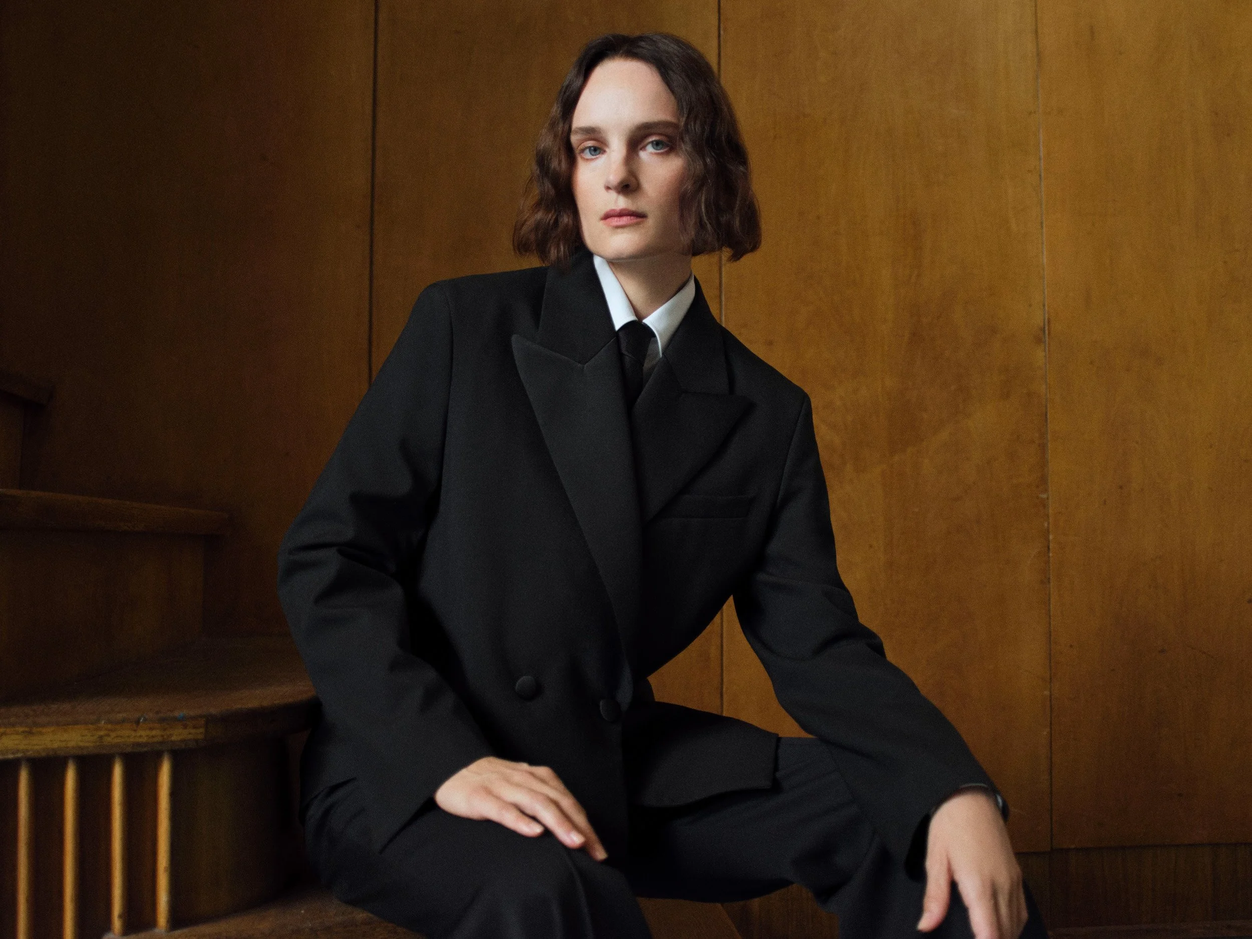 Person in an oversized black double-breasted suit with white shirt and tie, seated on wooden stairs against a wood-paneled wall.