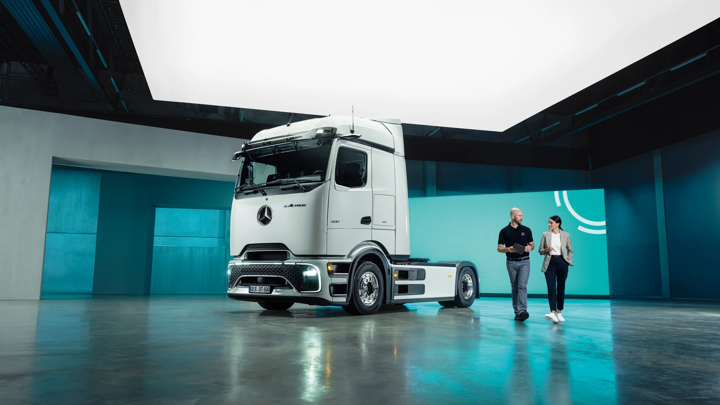 Two people walking and talking beside a white Mercedes-Benz eActros electric truck in a showroom with teal-lit walls.