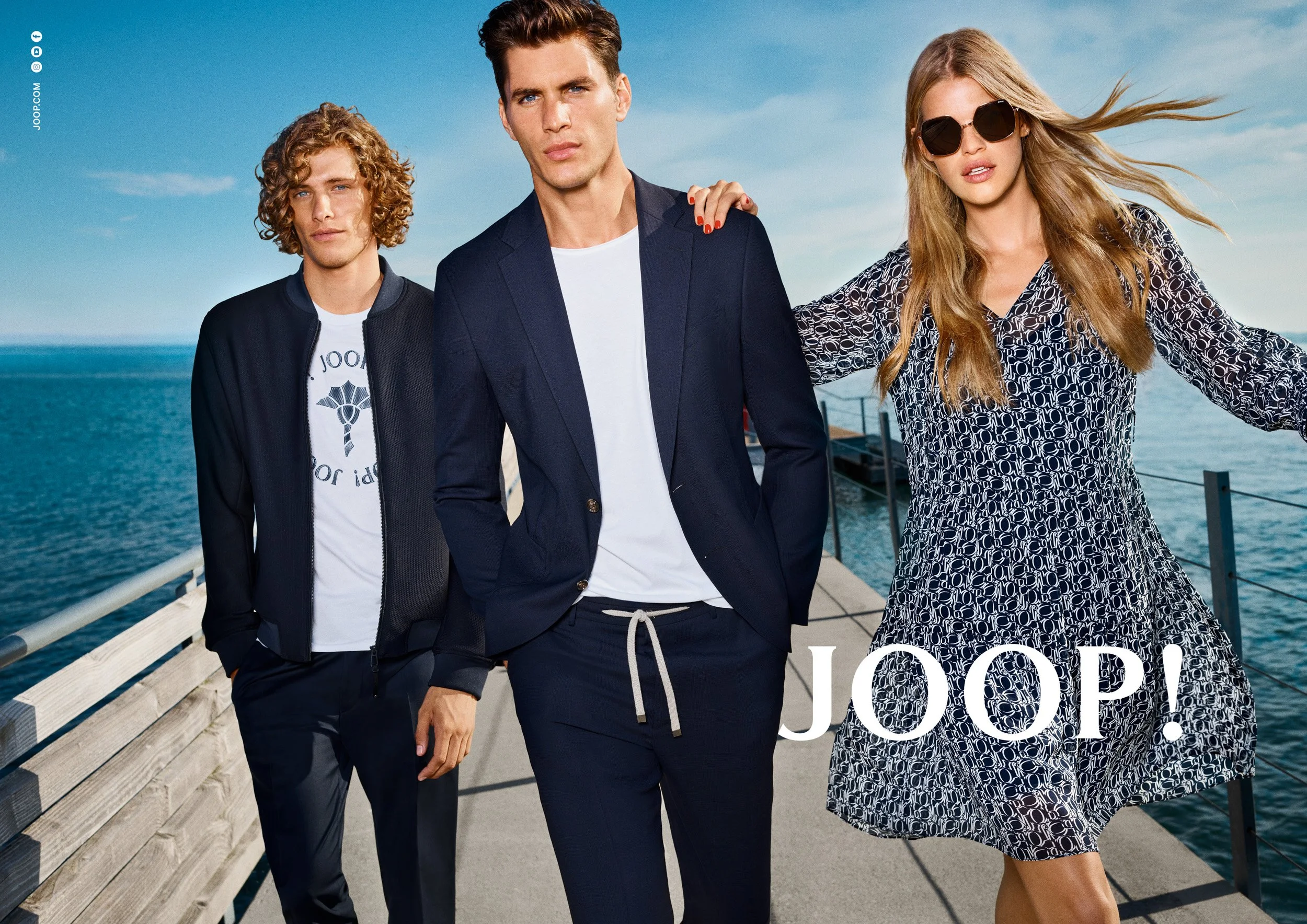 Two men in navy blazers and a woman in a printed navy dress walking along a wooden pier by the sea, with JOOP! logo.
