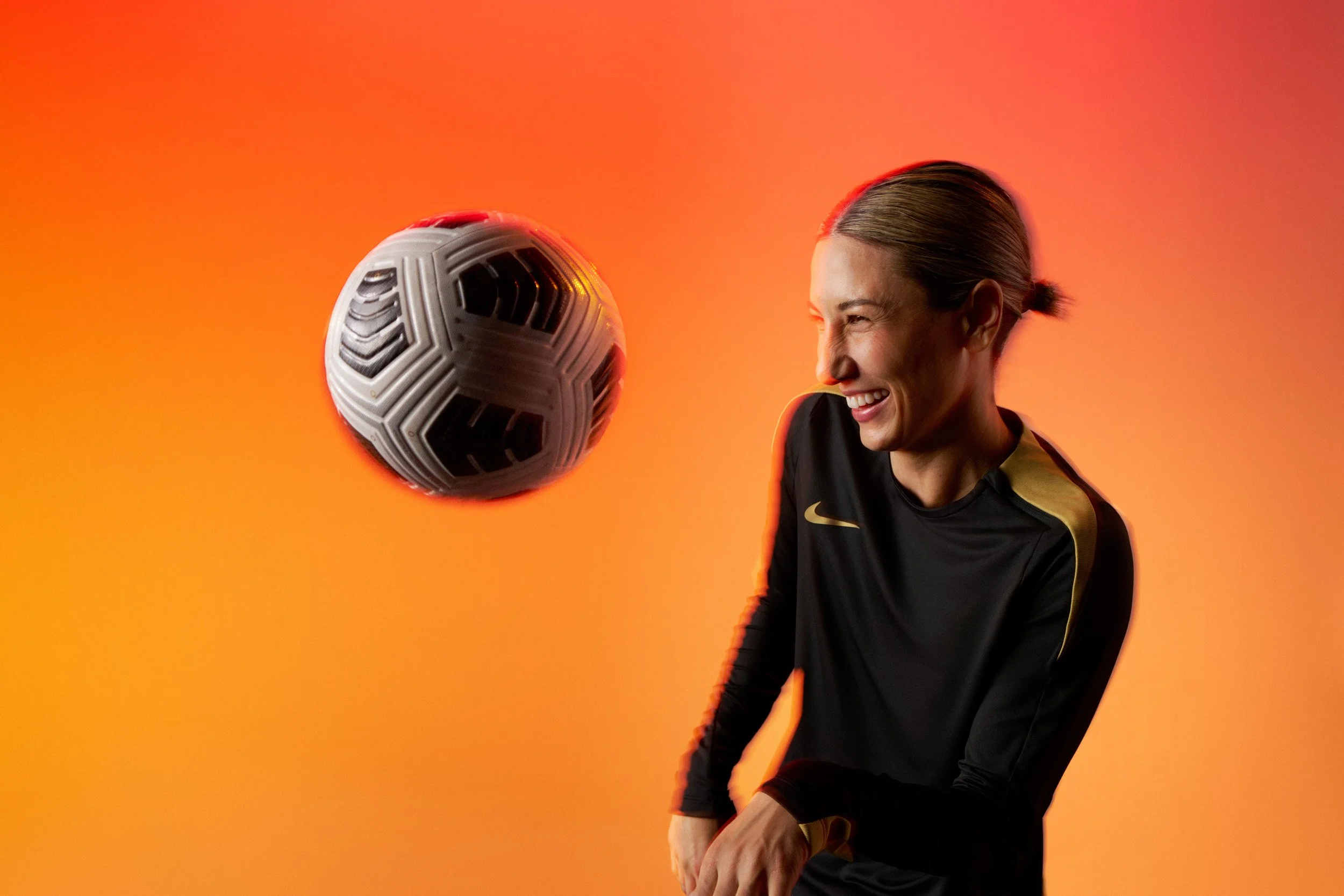Woman in a black and gold Nike top laughing while heading a soccer ball, against an orange gradient background.