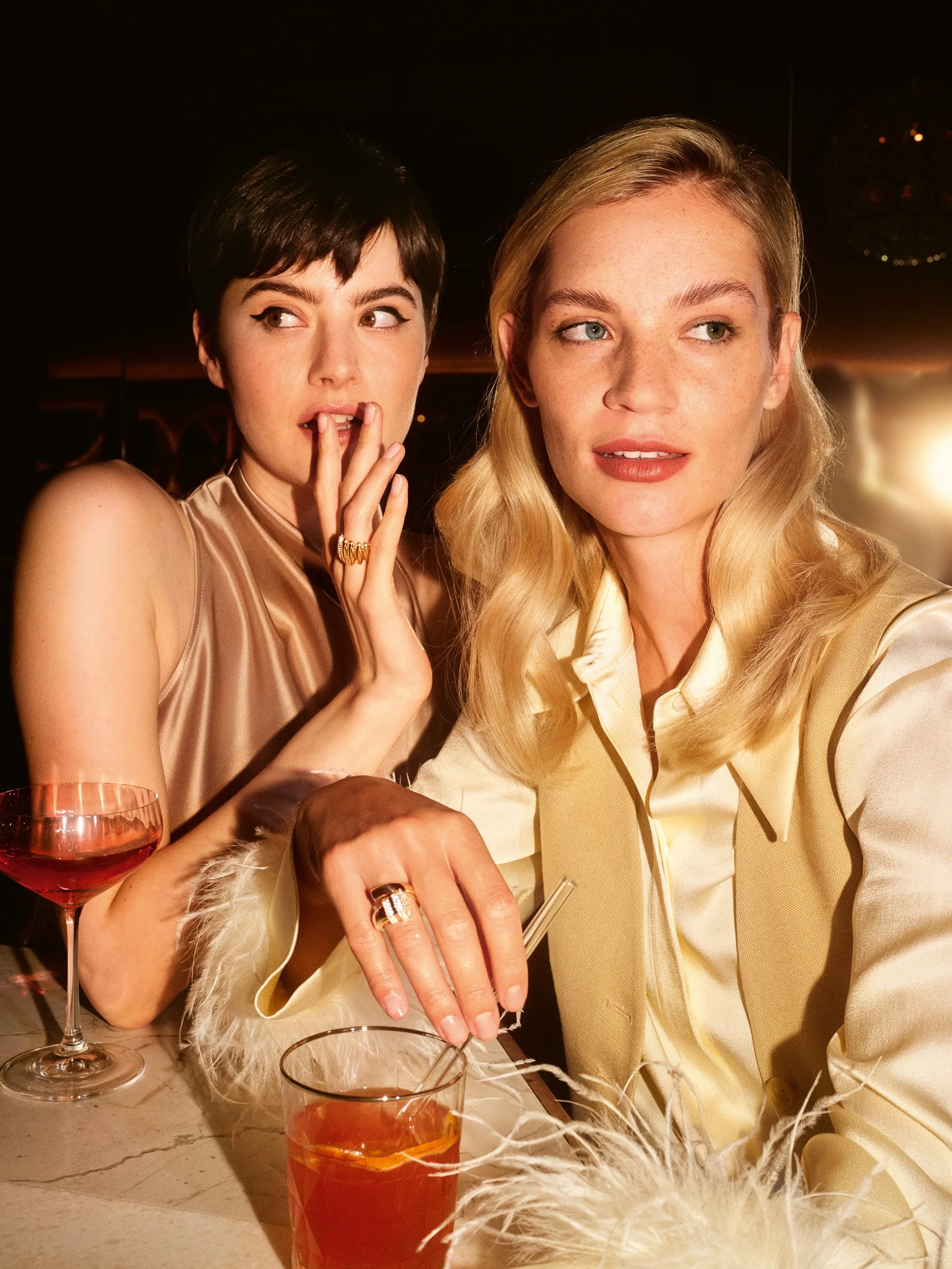 Two women at a bar in silky beige and cream tops, holding cocktail glasses under warm lighting.