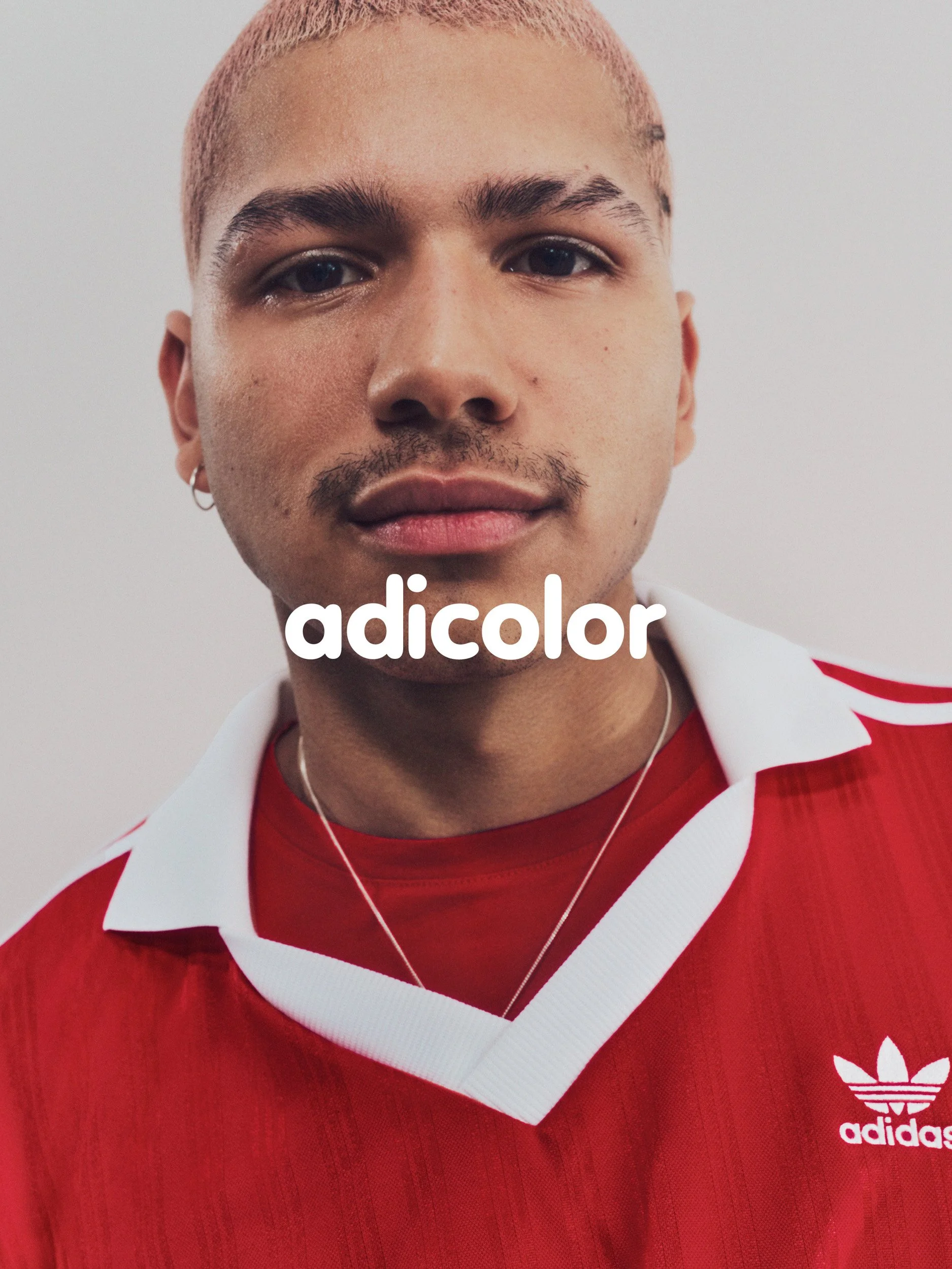 Close-up portrait of a man with pink hair wearing a red and white Adidas jersey, with "adicolor" text overlay.