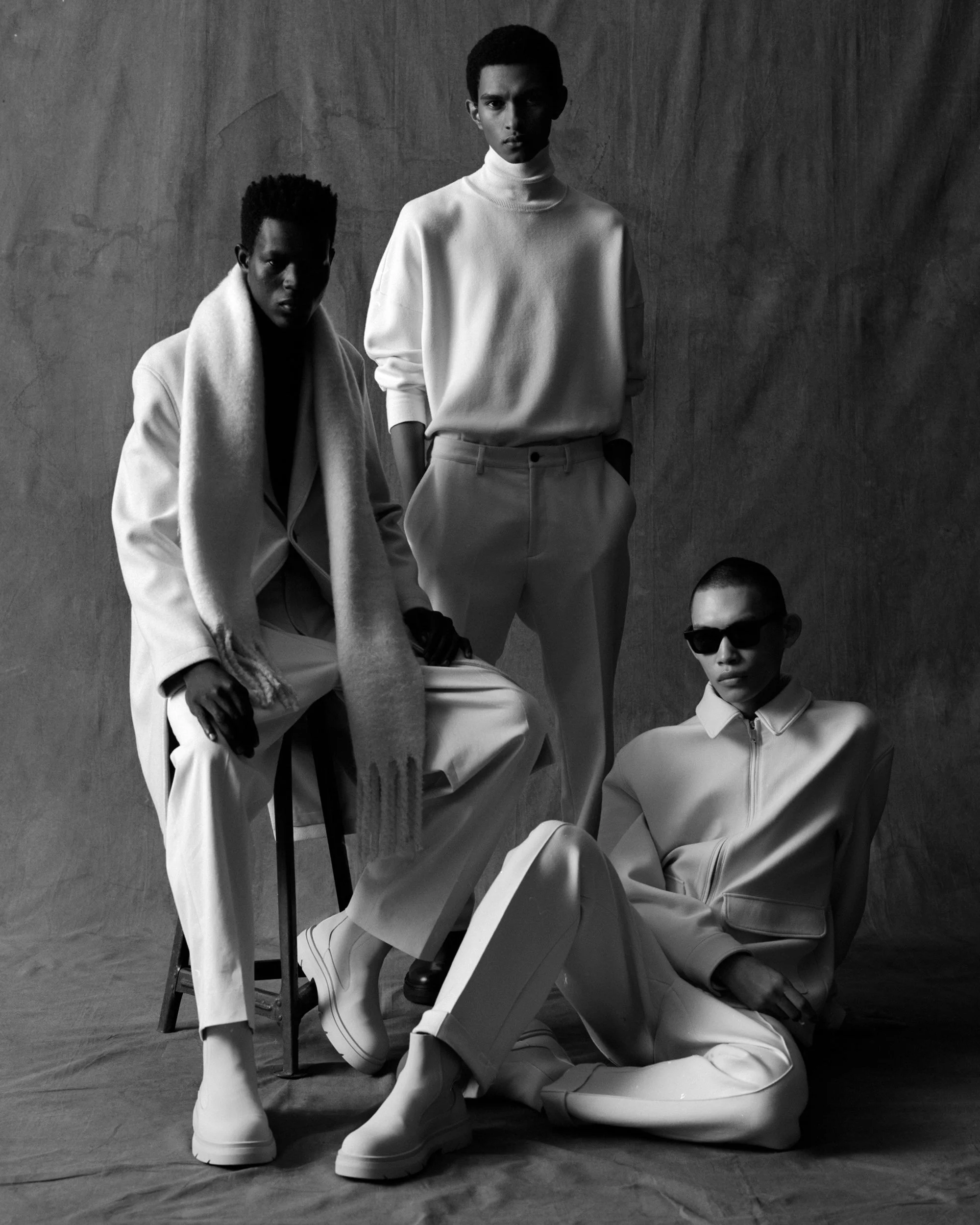 Black-and-white group portrait of three men in coordinated off-white tailored outerwear and turtlenecks.