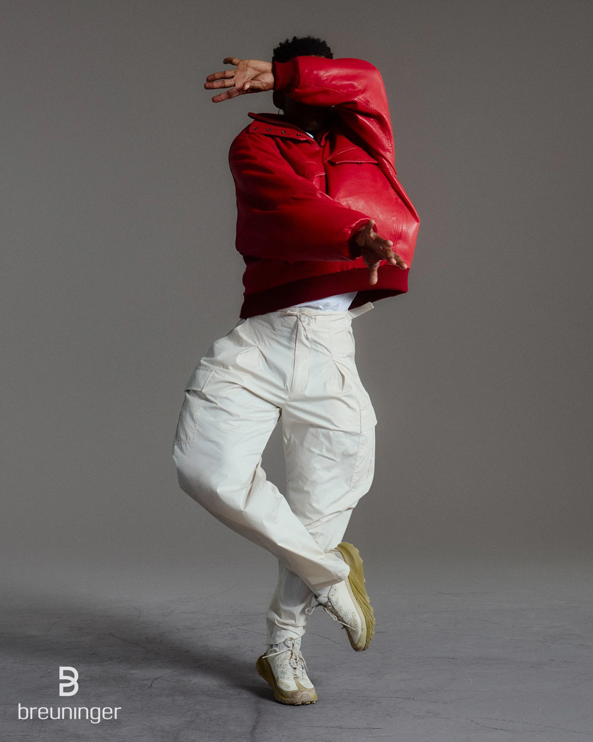 Man in a red leather jacket and cream cargo pants captured mid-motion against a gray background, with Breuninger logo.