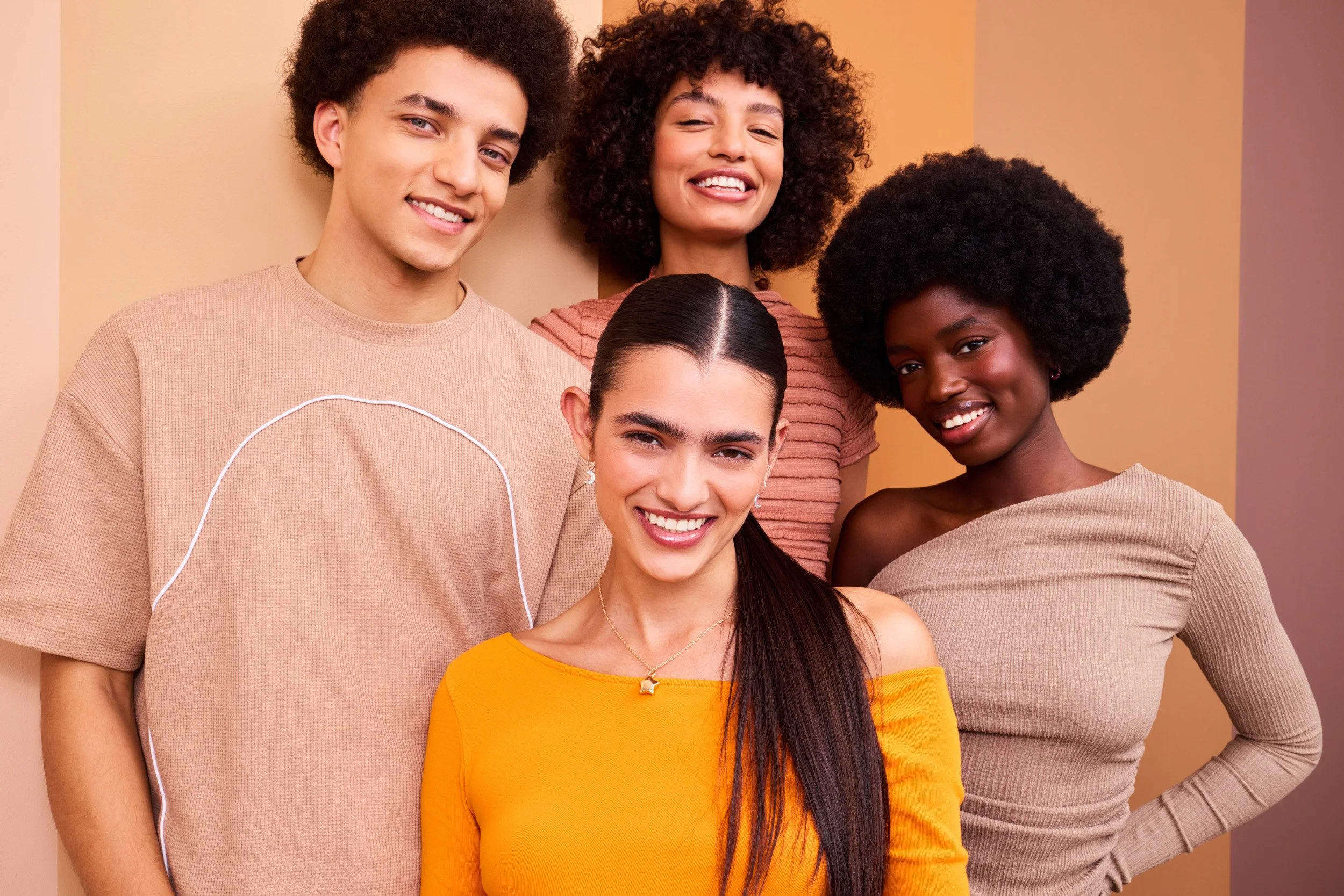 Group portrait of four people in warm-toned tan and orange tops, smiling against a striped beige and pink backdrop.