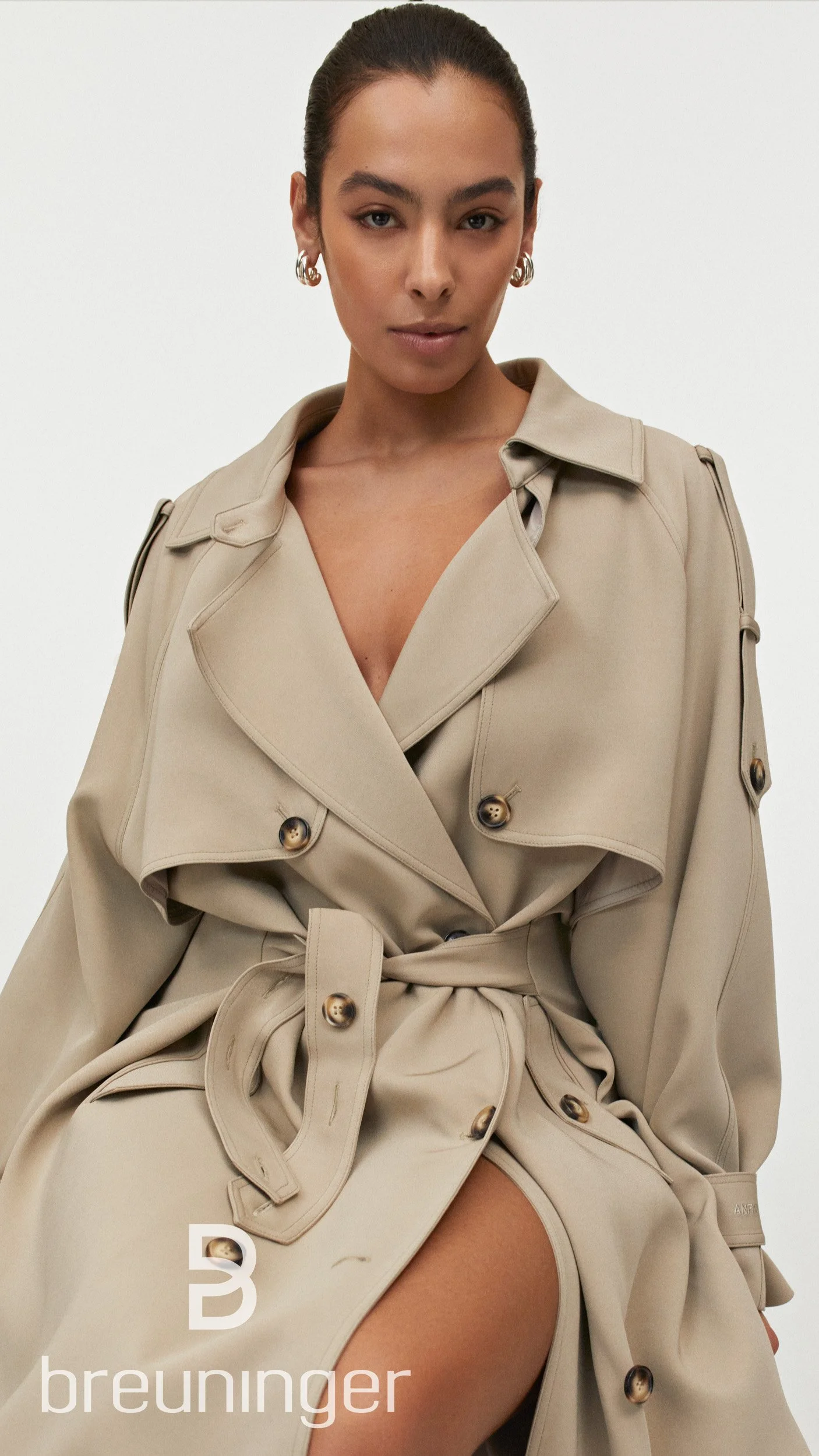 Close-up portrait of a woman in a beige trench coat with a plunging neckline, with Breuninger logo.