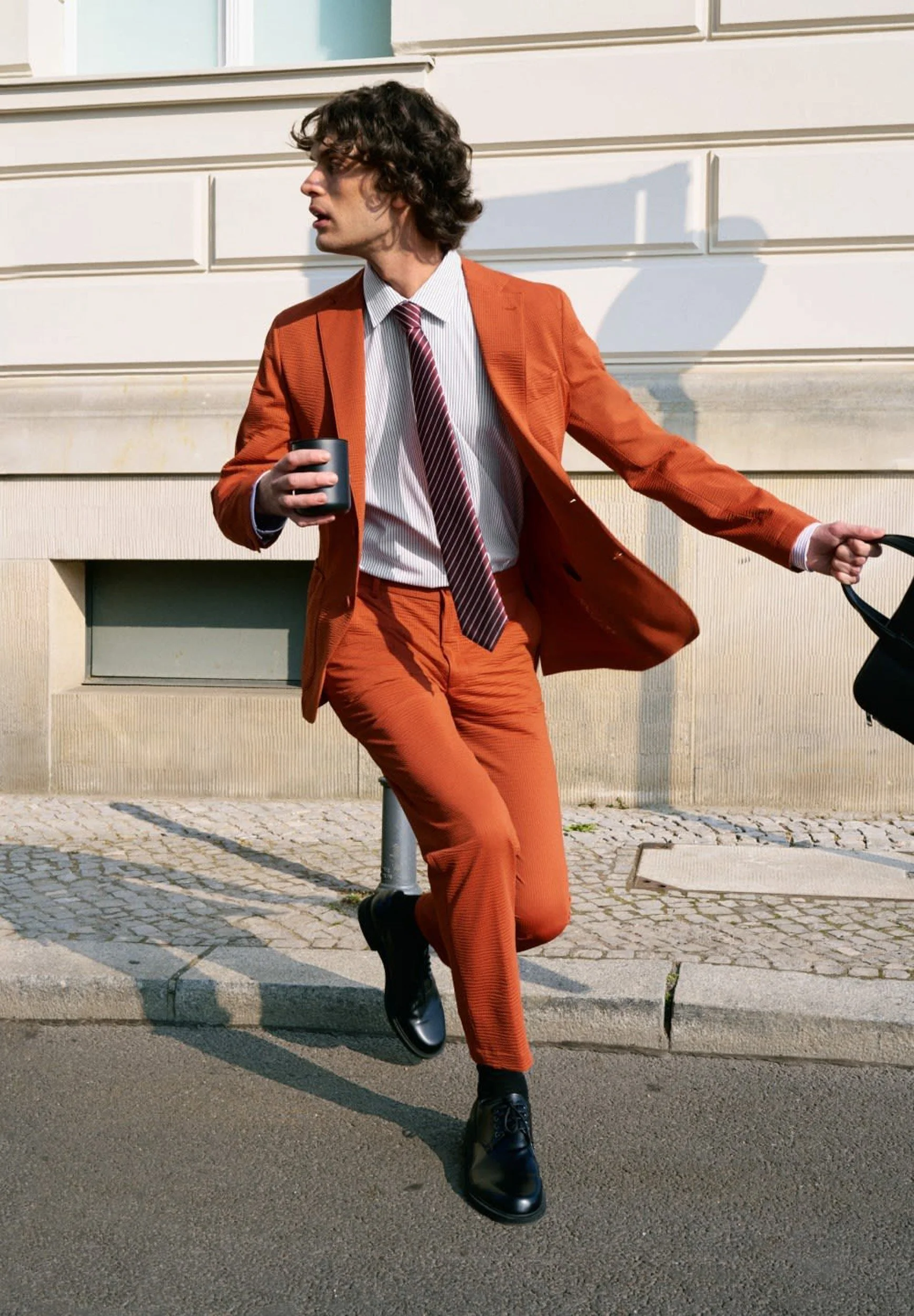 Man in a rust-orange suit and striped tie caught mid-stride on a cobblestone street, holding a coffee cup and bag.