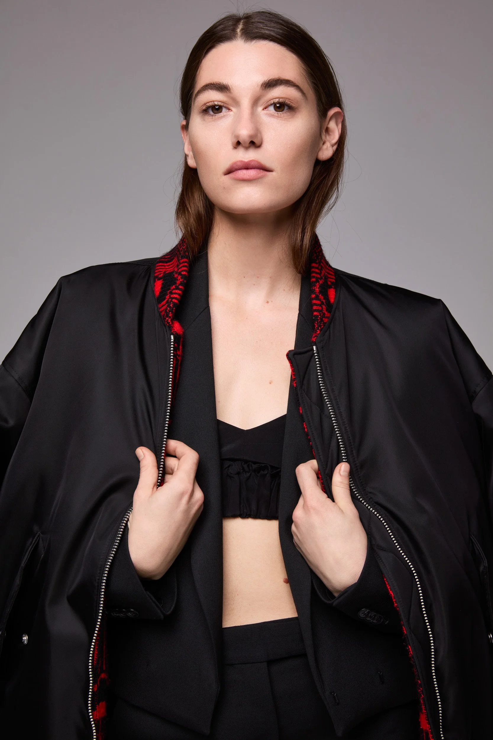 Close-up portrait of a woman in a black bomber jacket with red embroidered trim, unzipping it over a black cropped top.