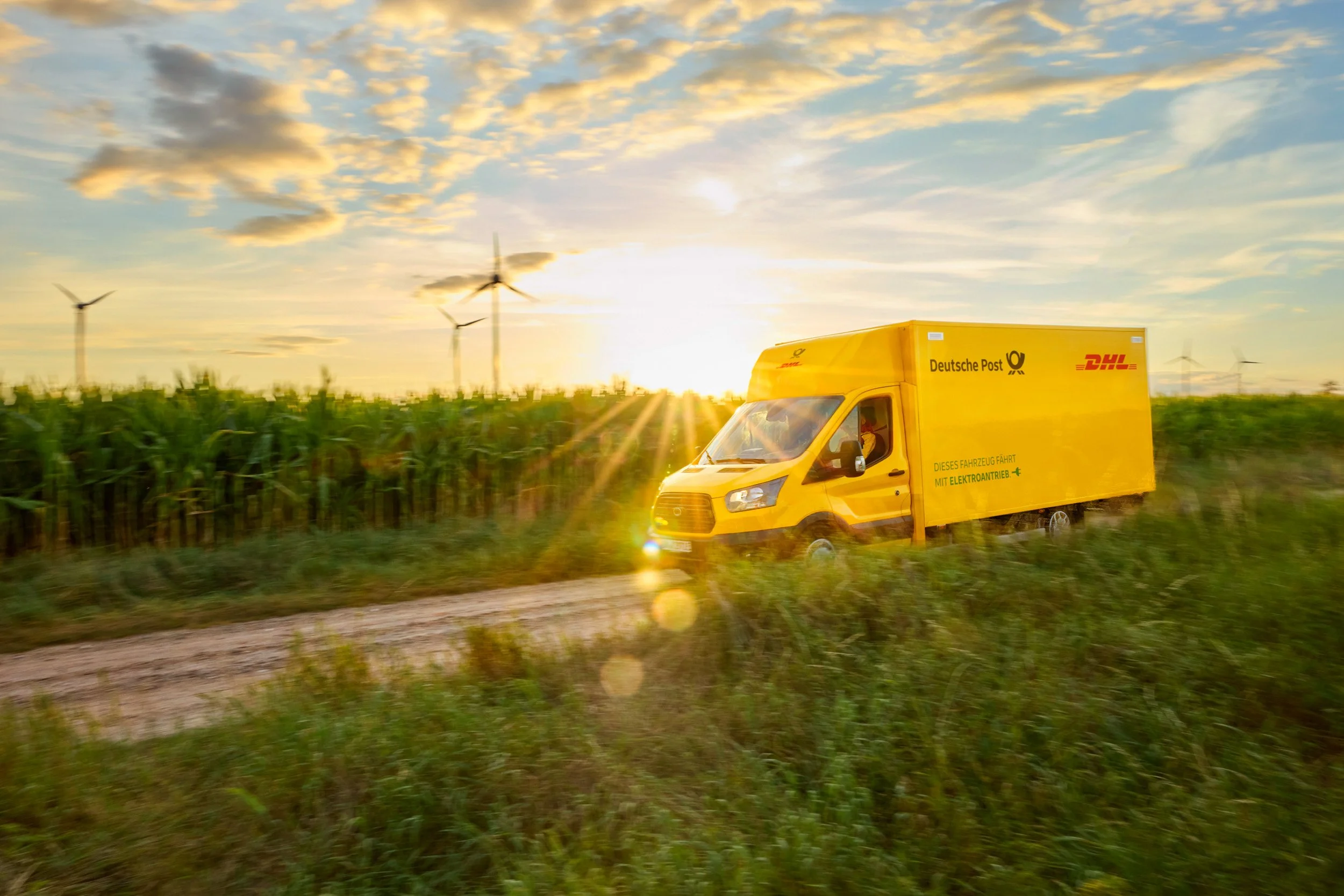 Yellow Deutsche Post DHL electric delivery van driving past a cornfield at sunset with wind turbines in the background.