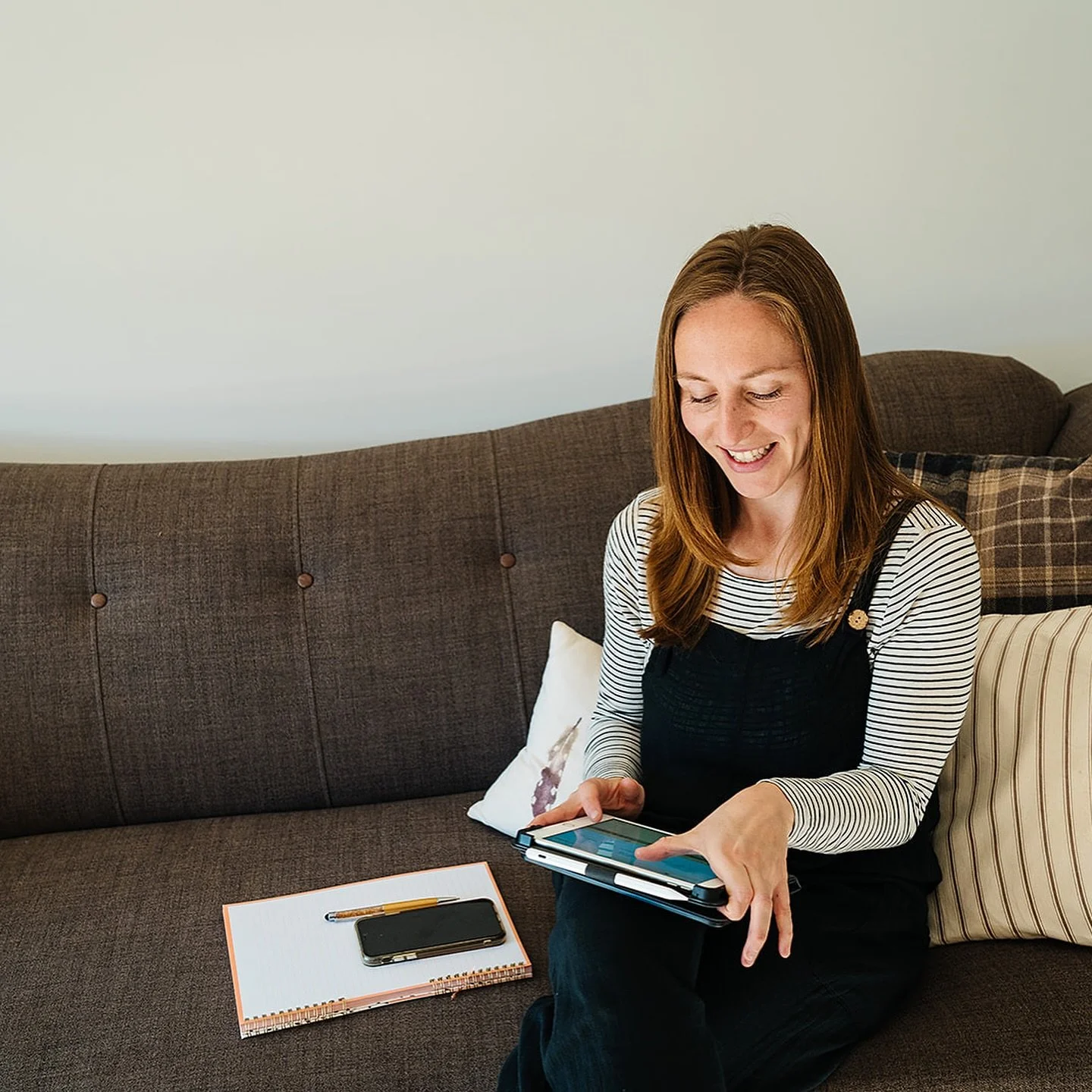 A woman with brown hair sitting on a brown couch, looking at a tablet and smiling. There is a notepad with a pen and smartphone on the table in front of her.