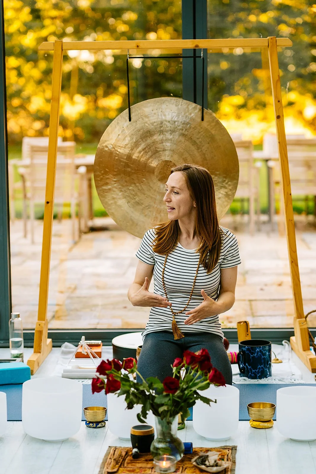 Woman sitting in front of a large gong, gesturing with her hands, gathered around with spiritual or meditation objects on a table, large windows showing trees with fall foliage in the background.