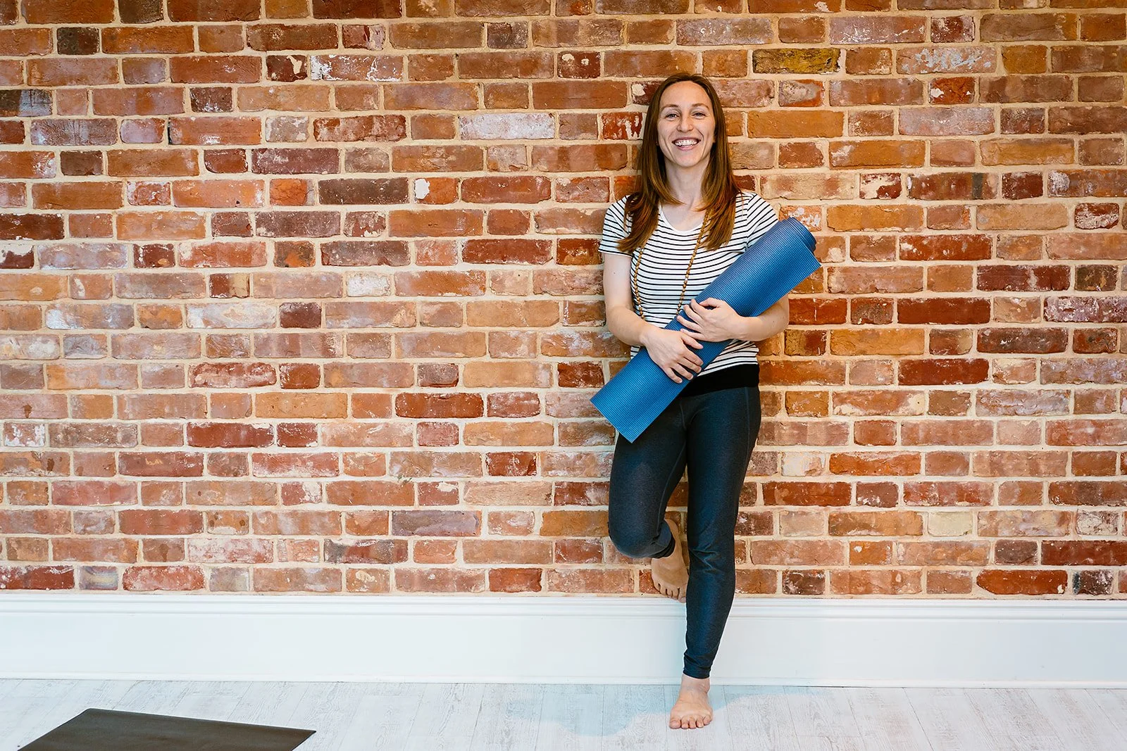 A woman with long brown hair standing barefoot in a yoga pose against a brick wall, holding a blue yoga mat.