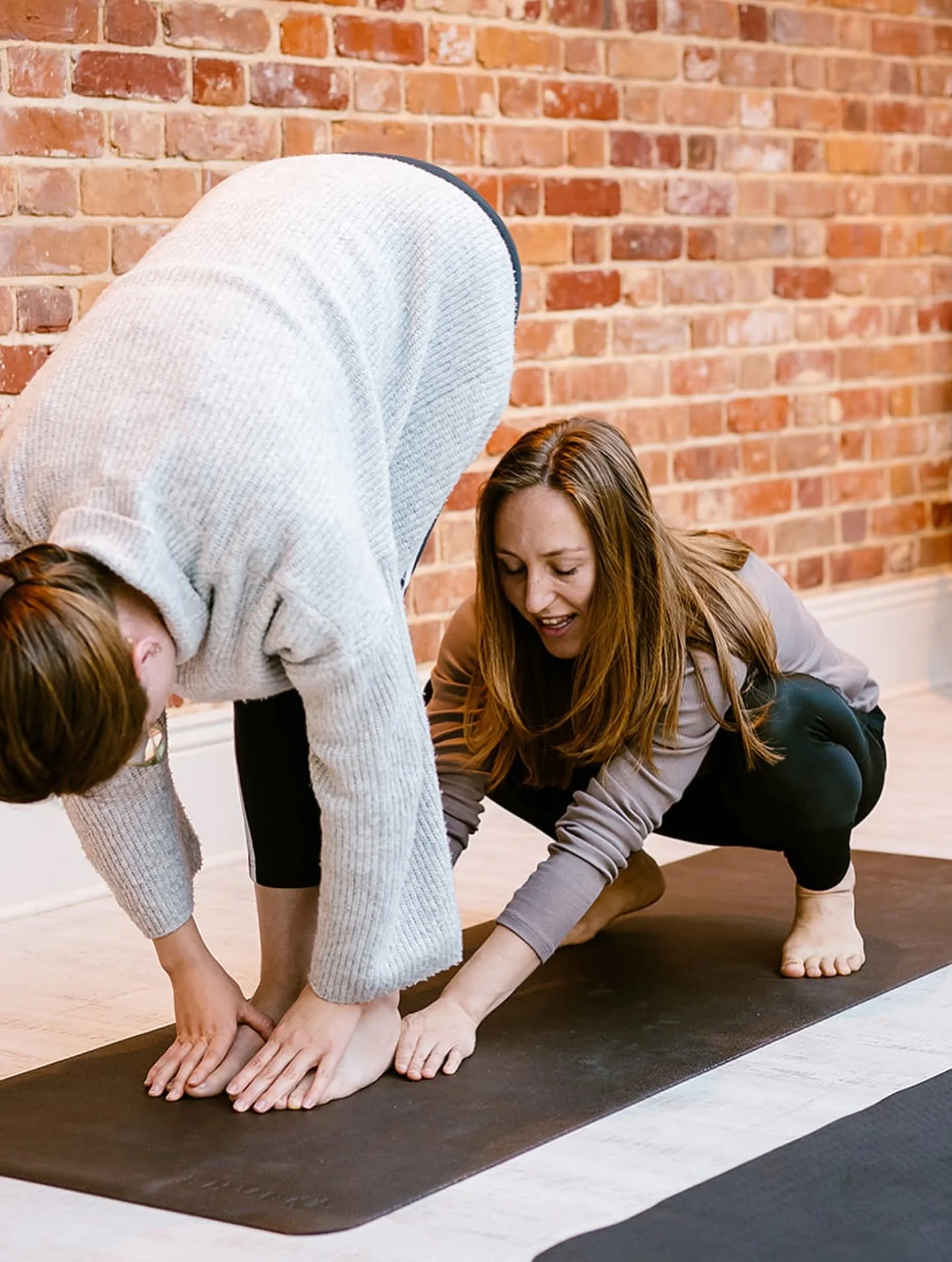 A woman and a child practicing yoga together on a black yoga mat against a brick wall, with the woman assisting the child in a forward bend pose.