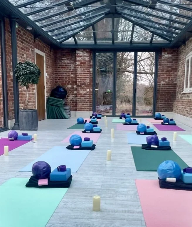 Indoor yoga studio with colorful mats, yoga blocks, bolsters, and candles arranged for a class, in a glass-enclosed room with brick walls and a view of trees outside.