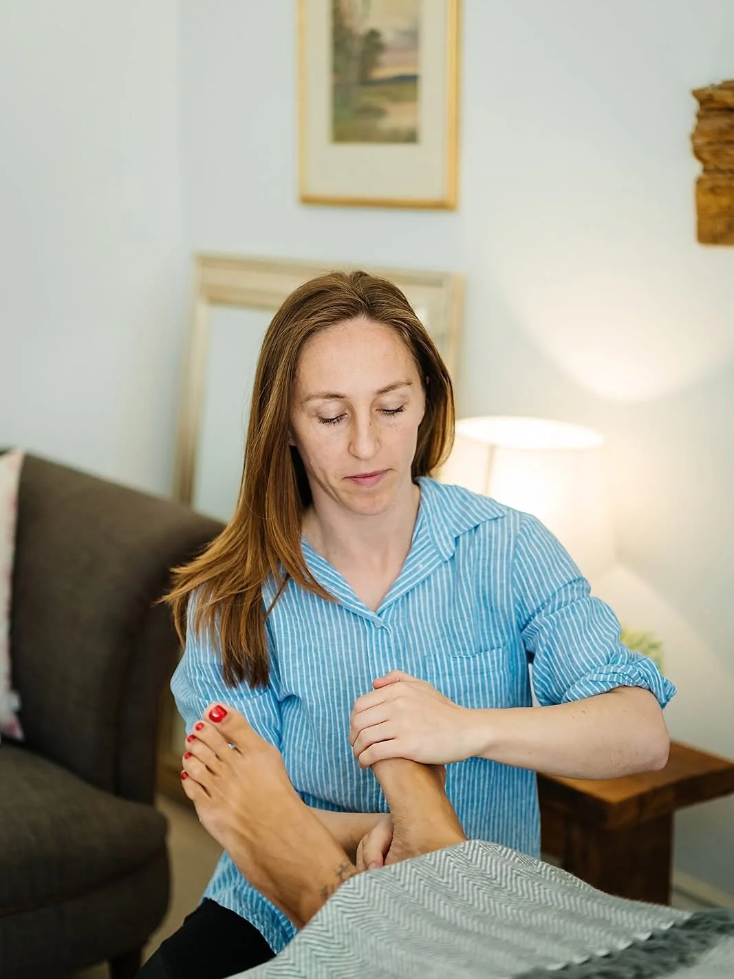 A woman in a blue striped shirt appears to be giving a foot massage or treatment to a person lying on a bed or table. The room has a lamp, framed picture, and wooden accents.
