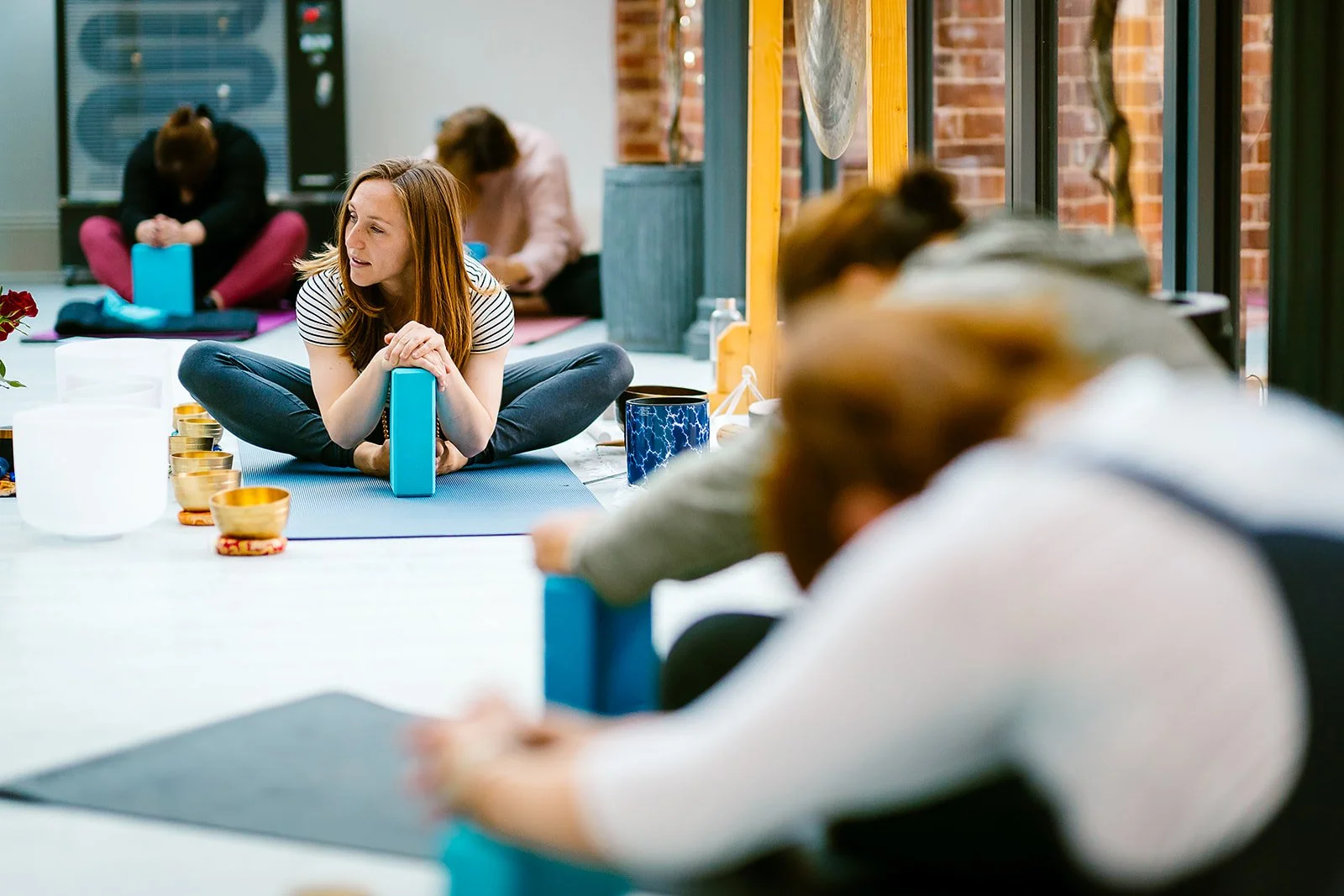 People participating in a seated yoga or meditation class in a bright room with large windows, brick walls, and natural light, with yoga mats, candles, and decorative items on the floor.