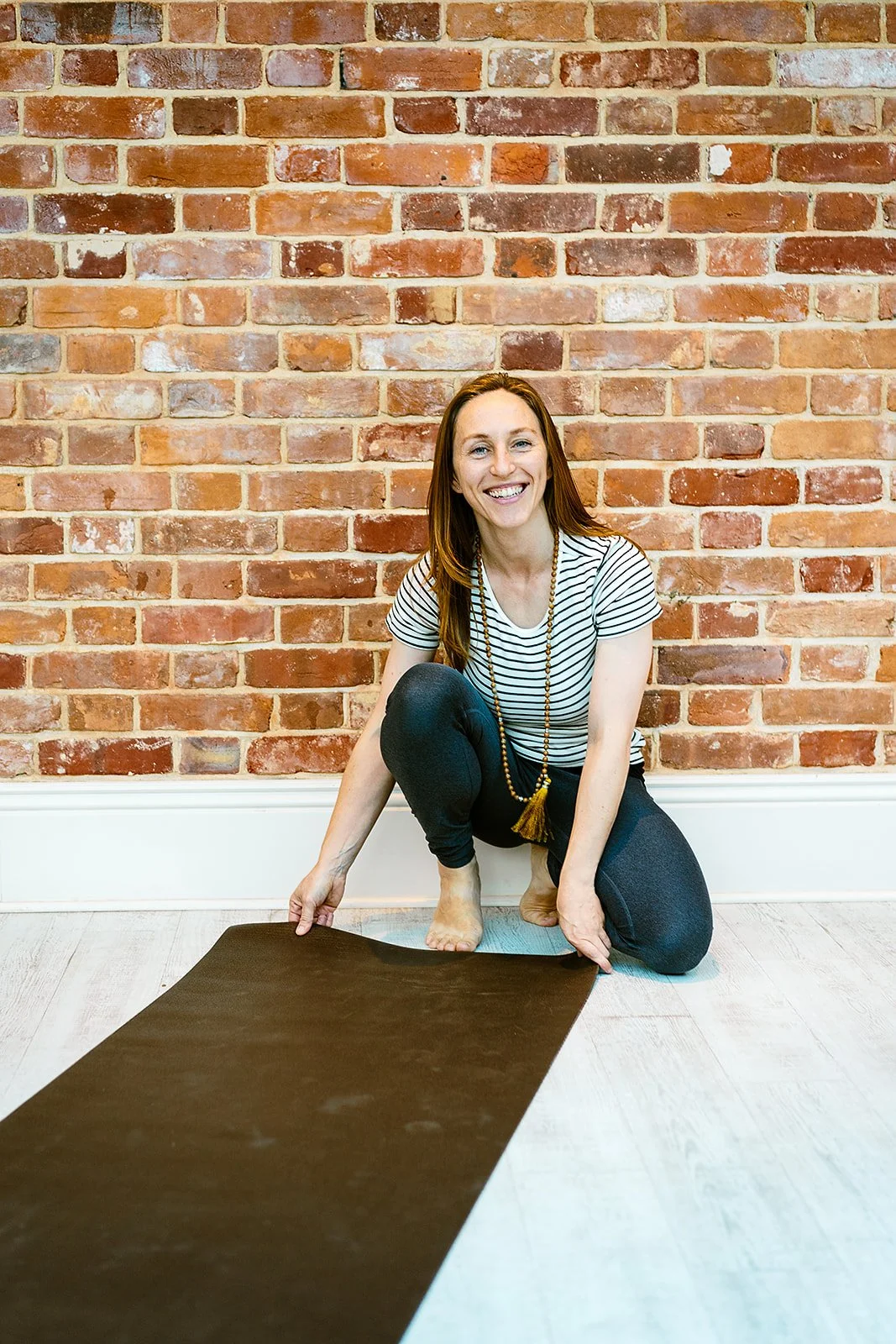 A woman with red hair and a striped shirt is sitting on the floor in front of a brick wall, smiling, with a yoga mat partially rolled out in front of her.