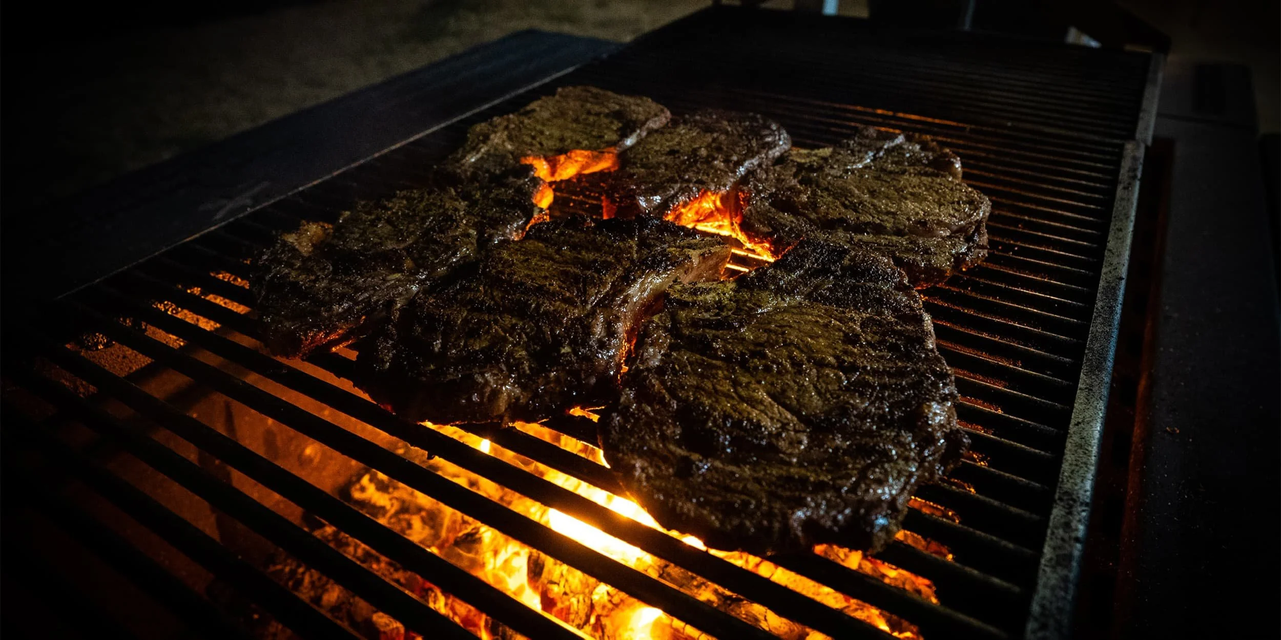 Steaks grilling on an outdoor barbecue at night with open flames underneath.