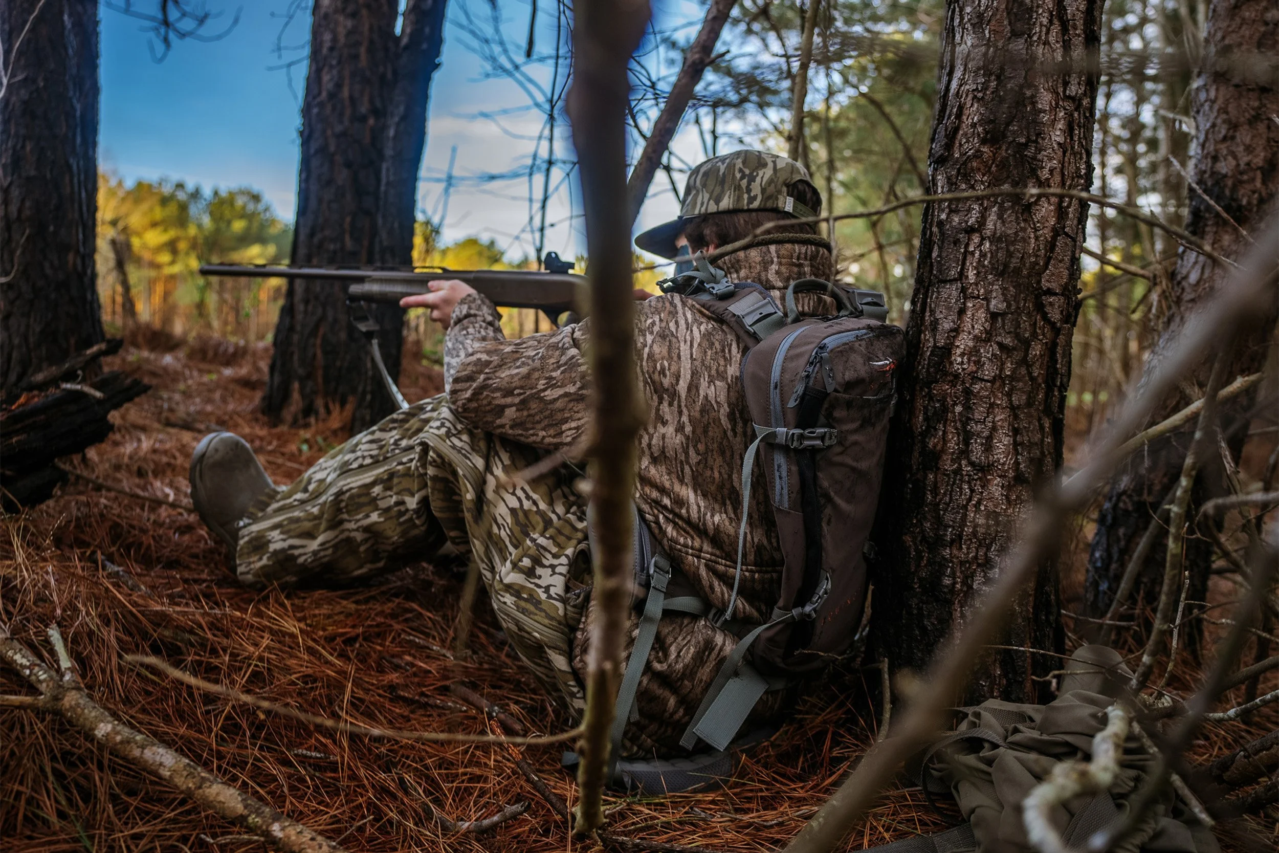 A person in camouflage clothing aiming a rifle while sitting among trees in a forest.