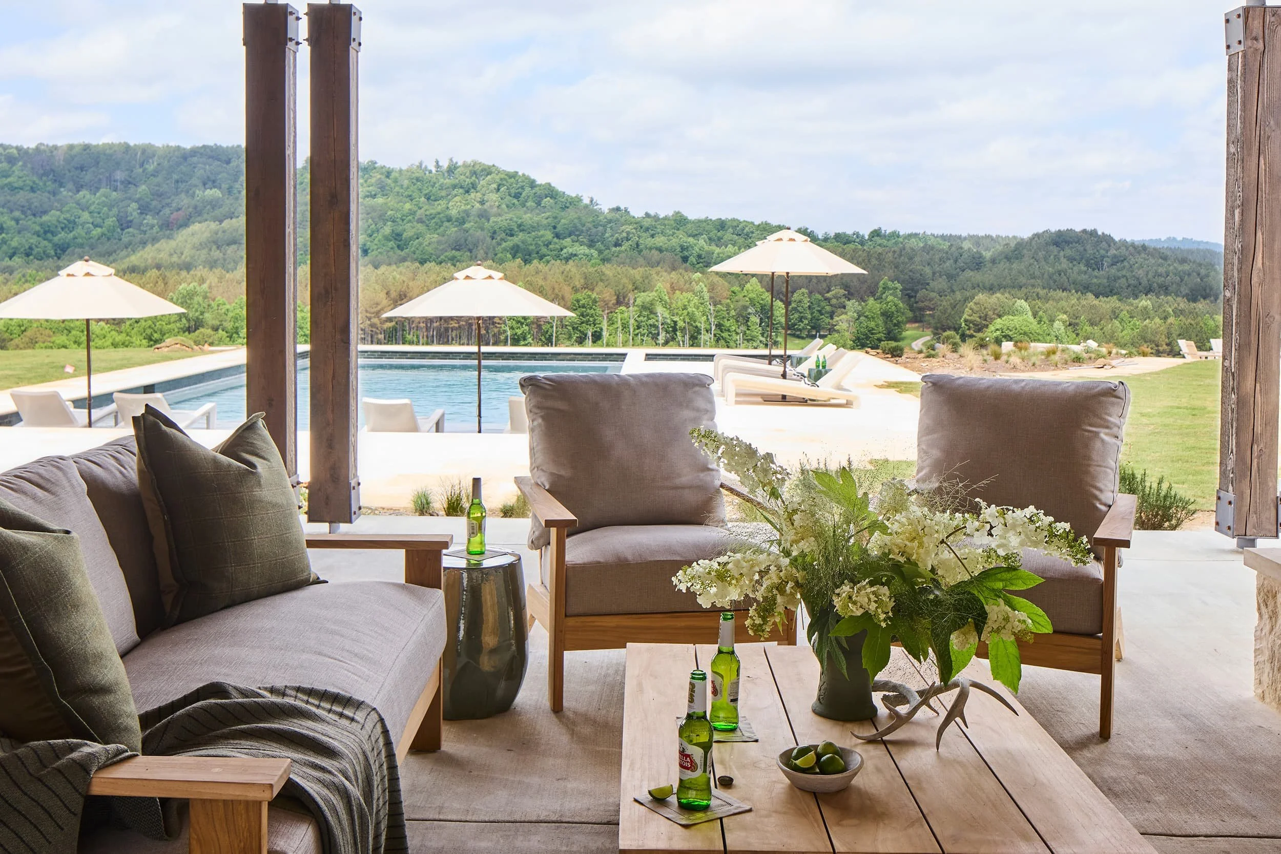 Interior view of a cozy outdoor lounge area overlooking a swimming pool and lush green mountains, decorated with sofas, pillows, a wooden coffee table with drinks and limes, and large potted flowers.
