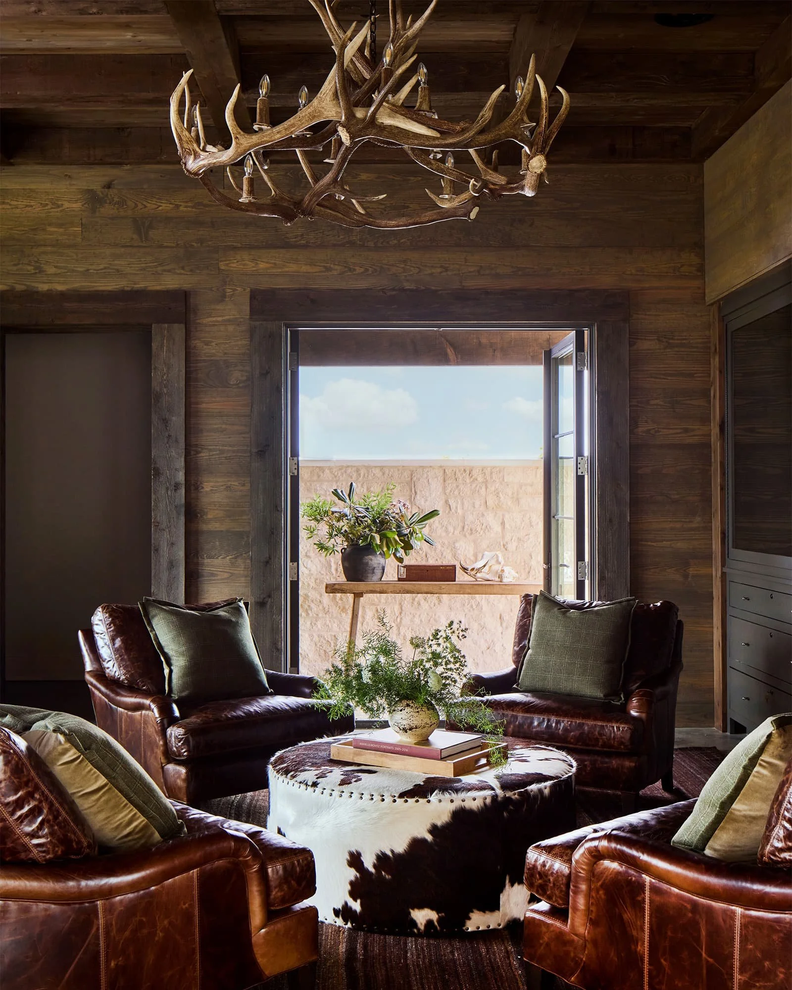 Cozy living room with leather armchairs, a cowhide ottoman, and a rustic chandelier made of antlers. Open door reveals a sunny patio with plants and a stone wall.