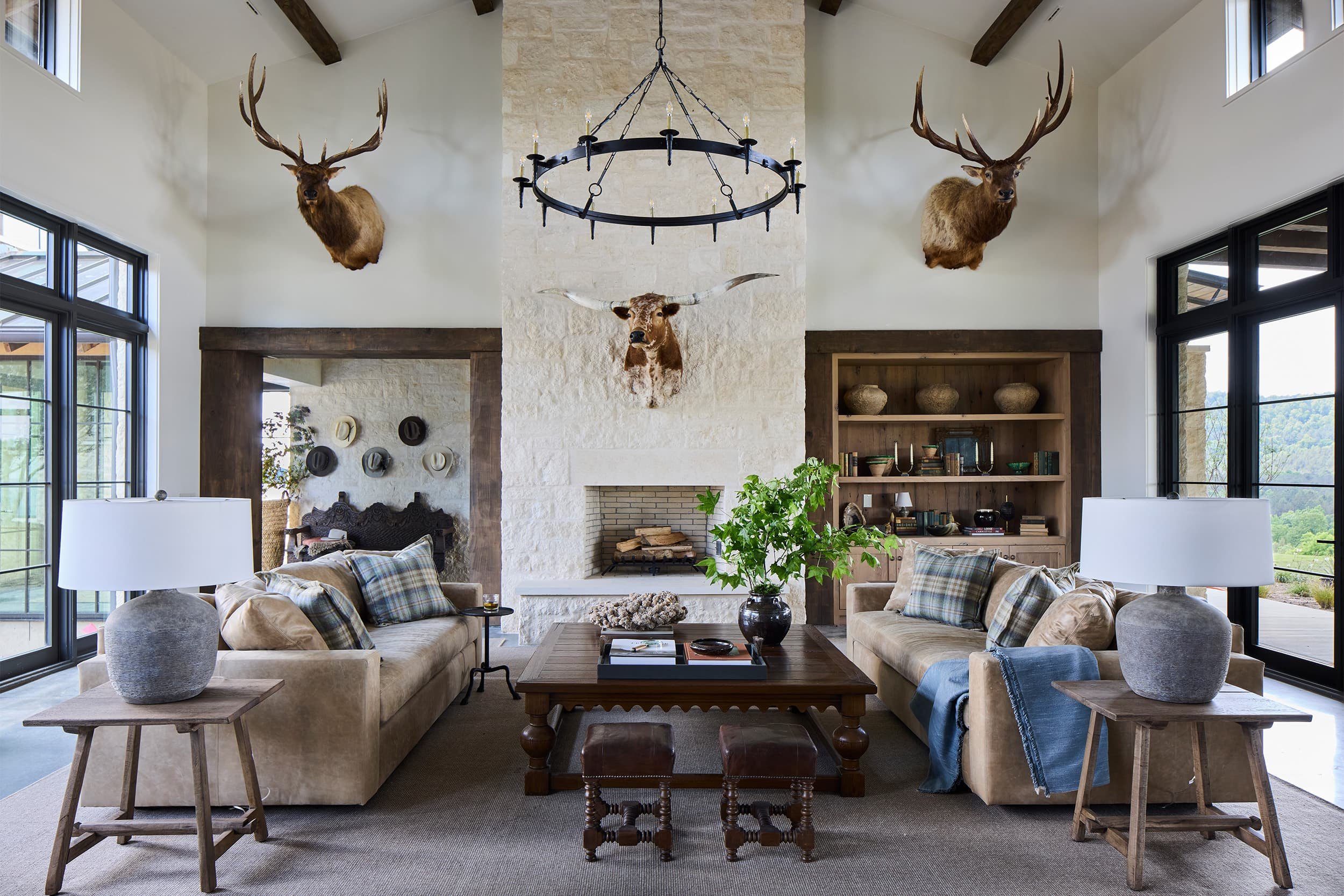 Living room with beige sofas, plaid pillows, wooden coffee table, large windows, and mounted deer heads with antlers on the walls.