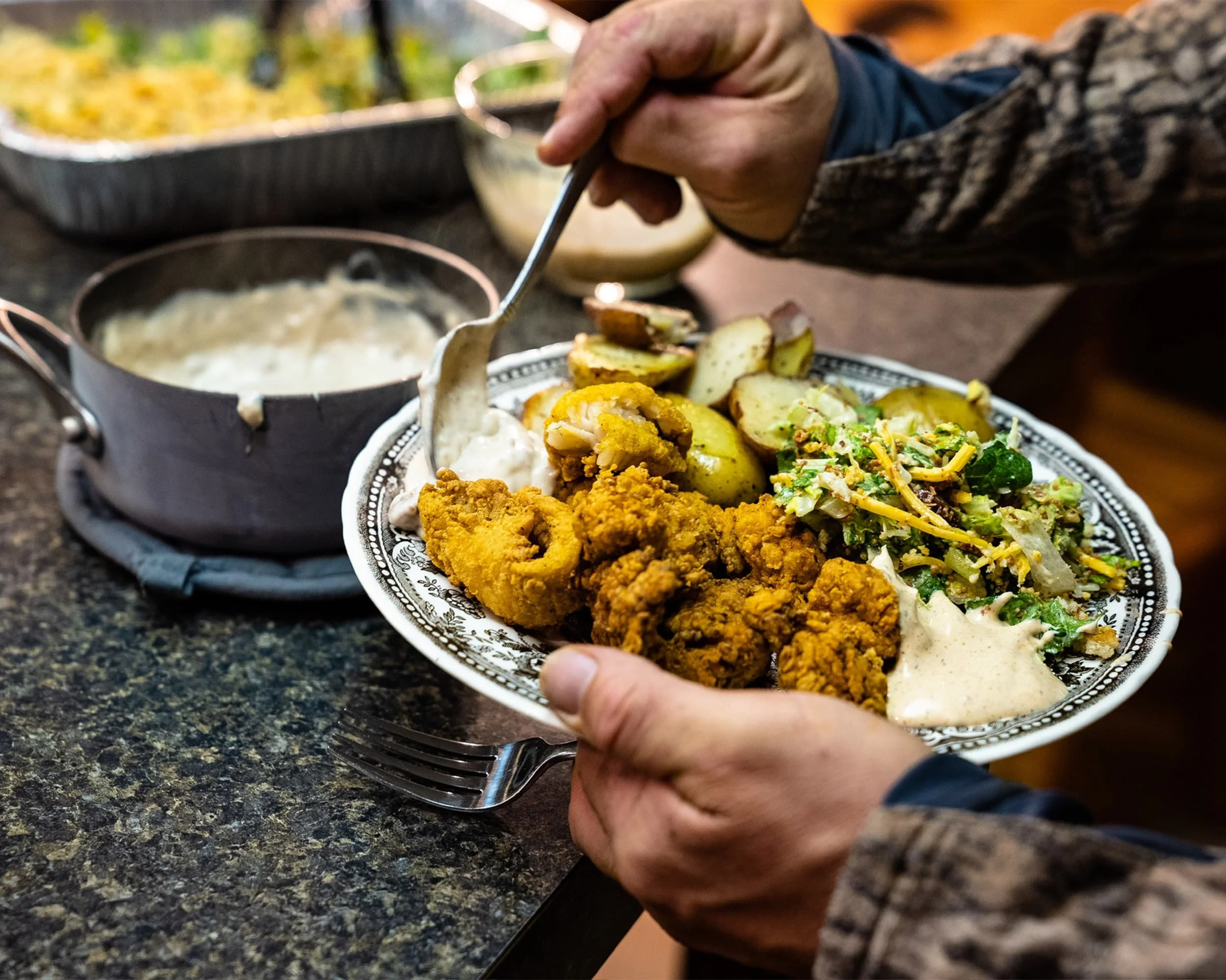 A person holding a platter of fried chicken, potato wedges, salad, and a creamy dip, with a pot of gravy and a dish of mixed vegetables in the background.