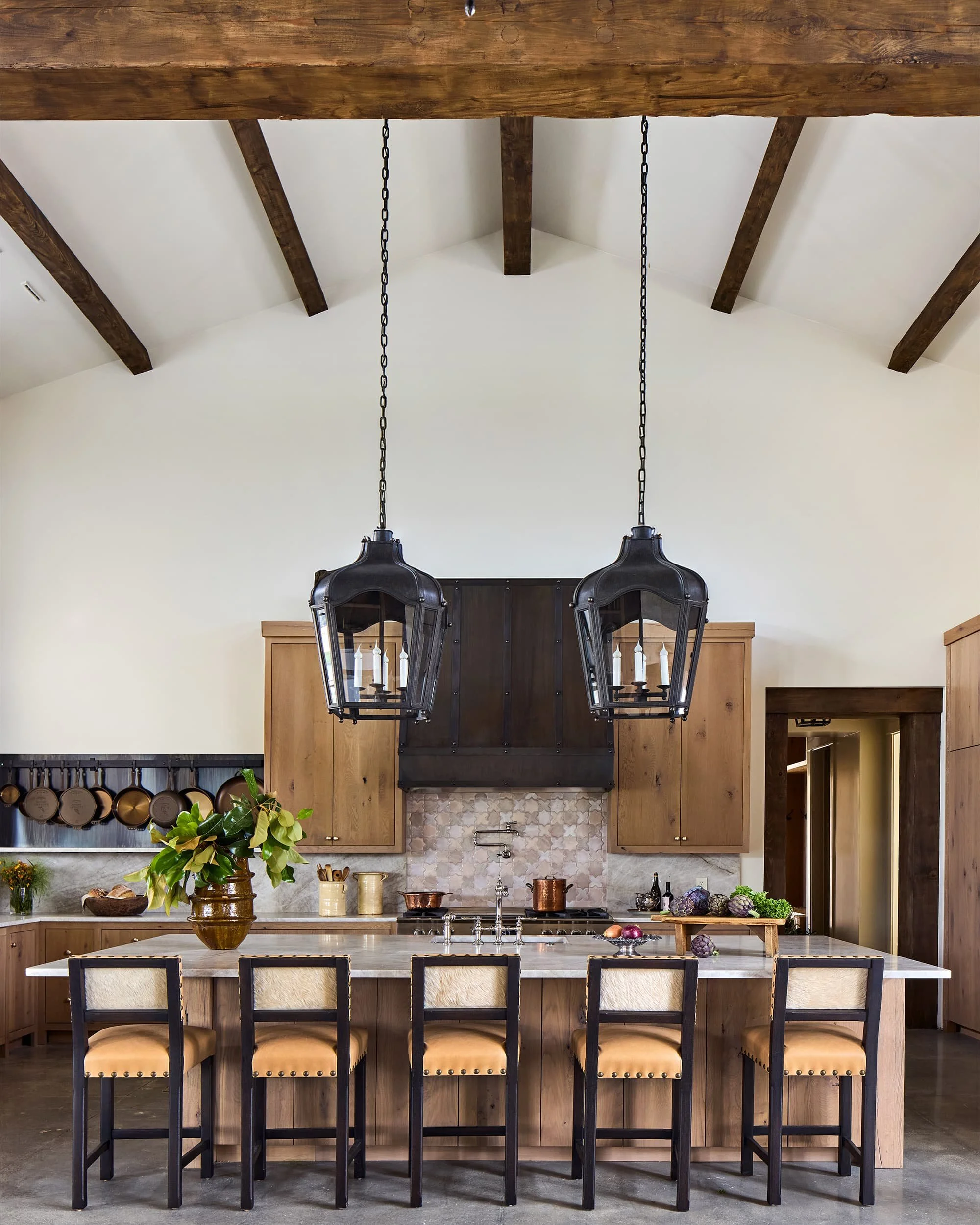 A rustic kitchen with a large white island countertop, six black high-back chairs with beige cushions, wooden cabinets, and a modern black and glass chandelier hanging from a vaulted wooden ceiling.