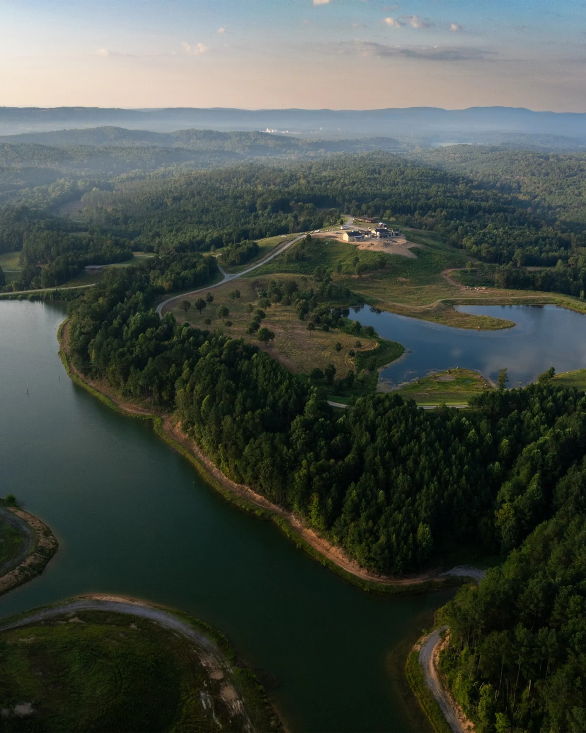 Aerial view of a large river winding through lush green forests and rolling hills with a few buildings on a hilltop. Distant mountain range with mist and scattered clouds in the sky.