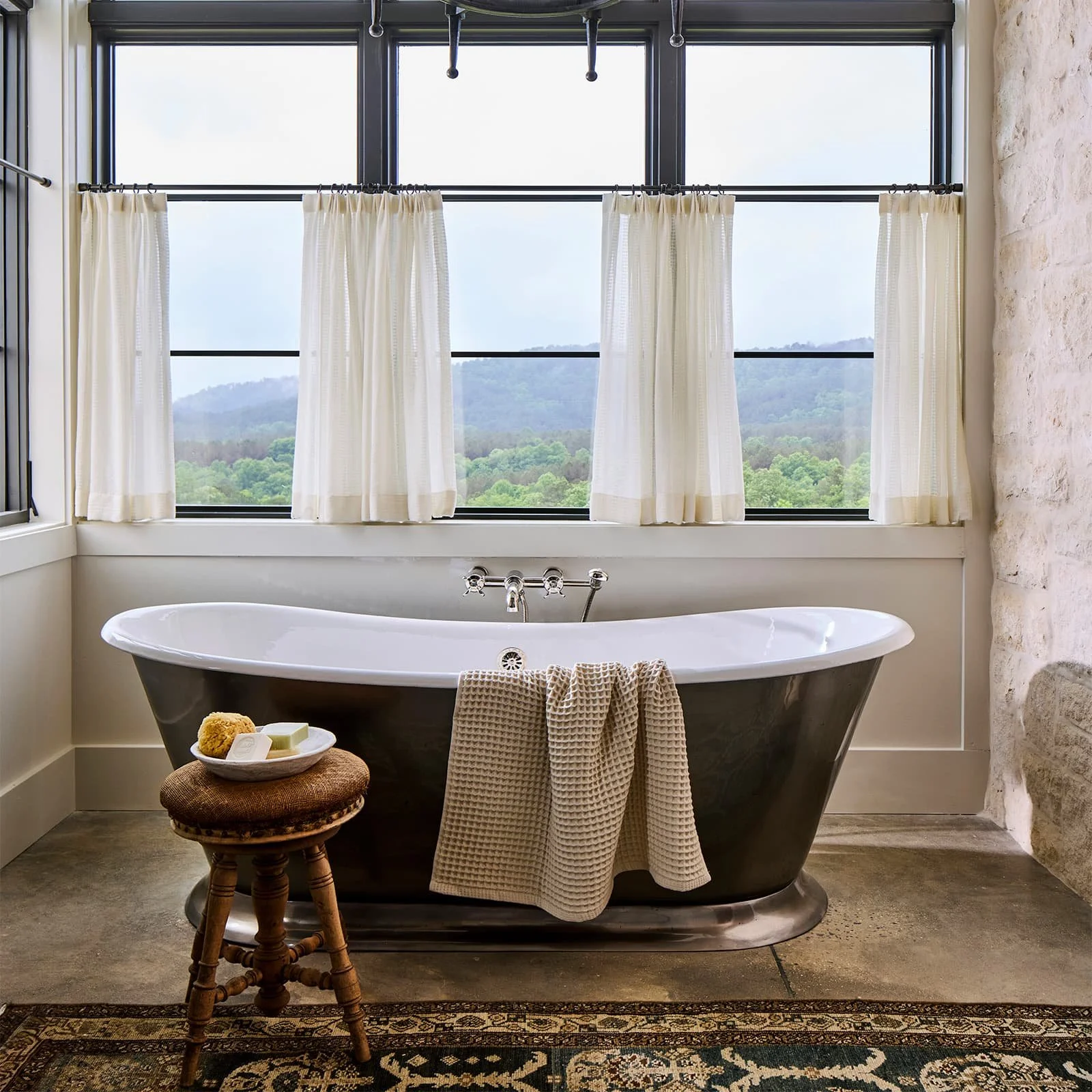 A vintage-style bathroom with a black clawfoot bathtub, a small wooden stool with soap and a bath sponge on it, and a large window with white curtains showing a scenic mountain view in the background.