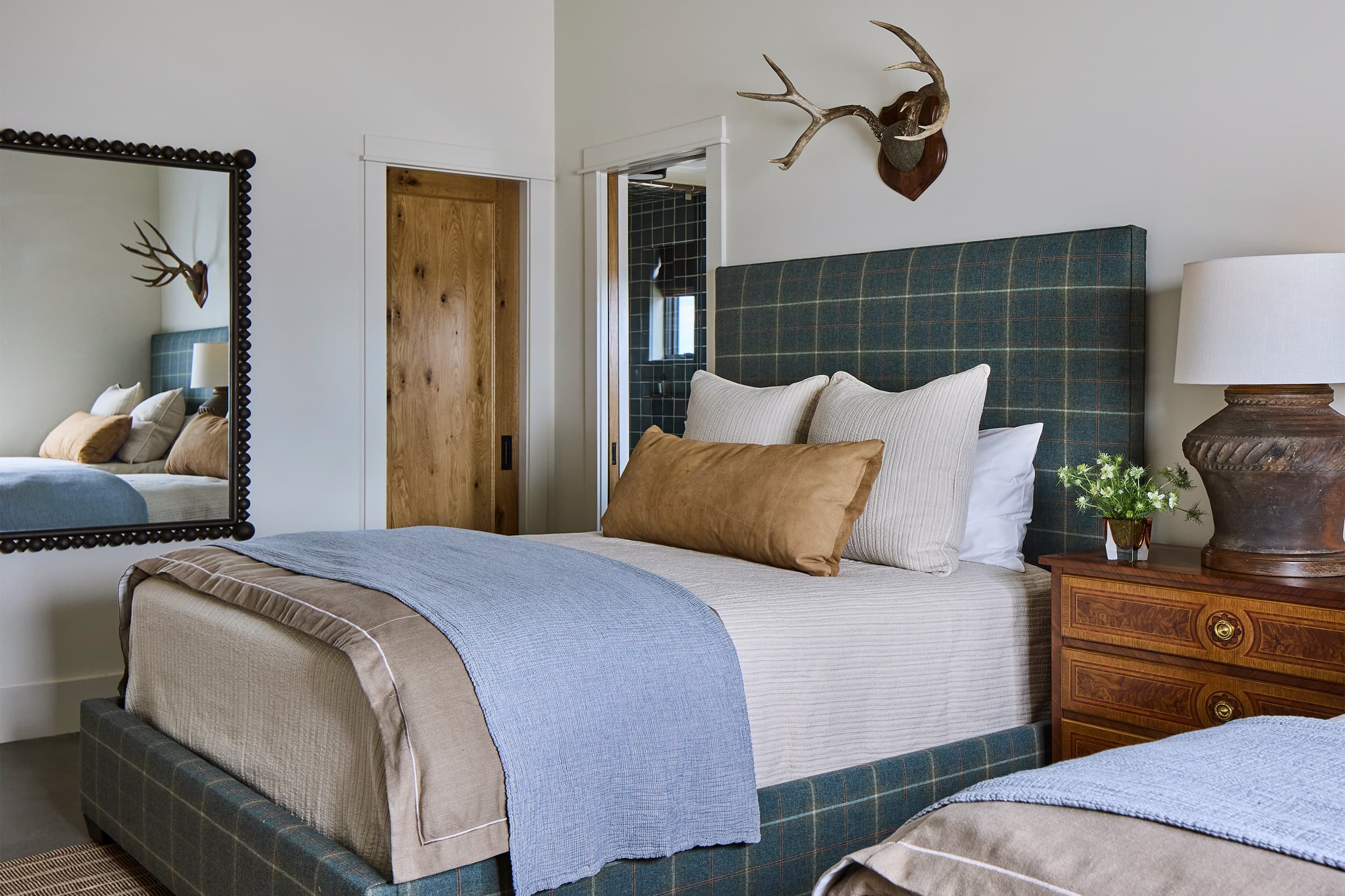 Bedroom with a bed that has a plaid upholstered headboard, pillows, and a beige blanket, with a wooden nightstand, a lamp, a mirror with a beaded frame, and wall-mounted antlers.