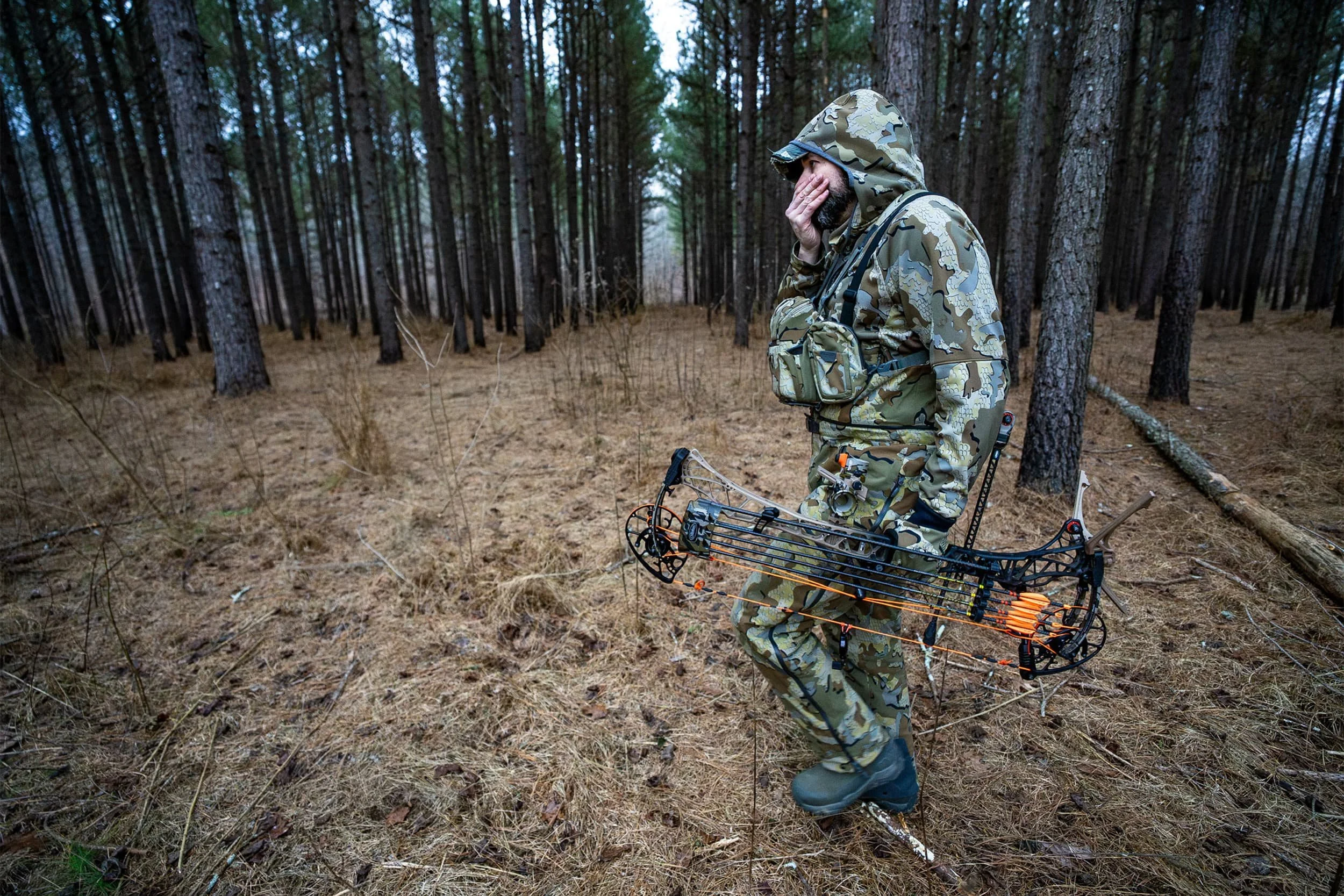A man in camouflage clothing stands in a forest, holding a compound bow with an arrow loaded, appearing to be preparing for archery.