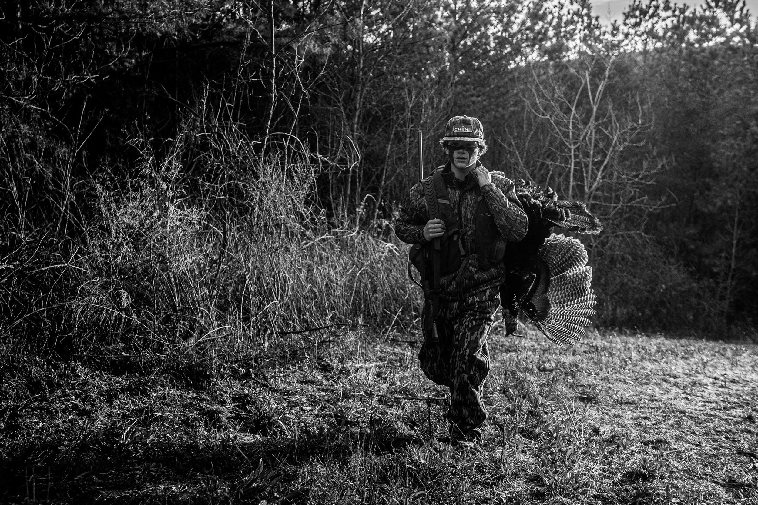 A person dressed in camouflage clothing carrying a rifle over their shoulder and a turkey in their hand, walking through a forested area.